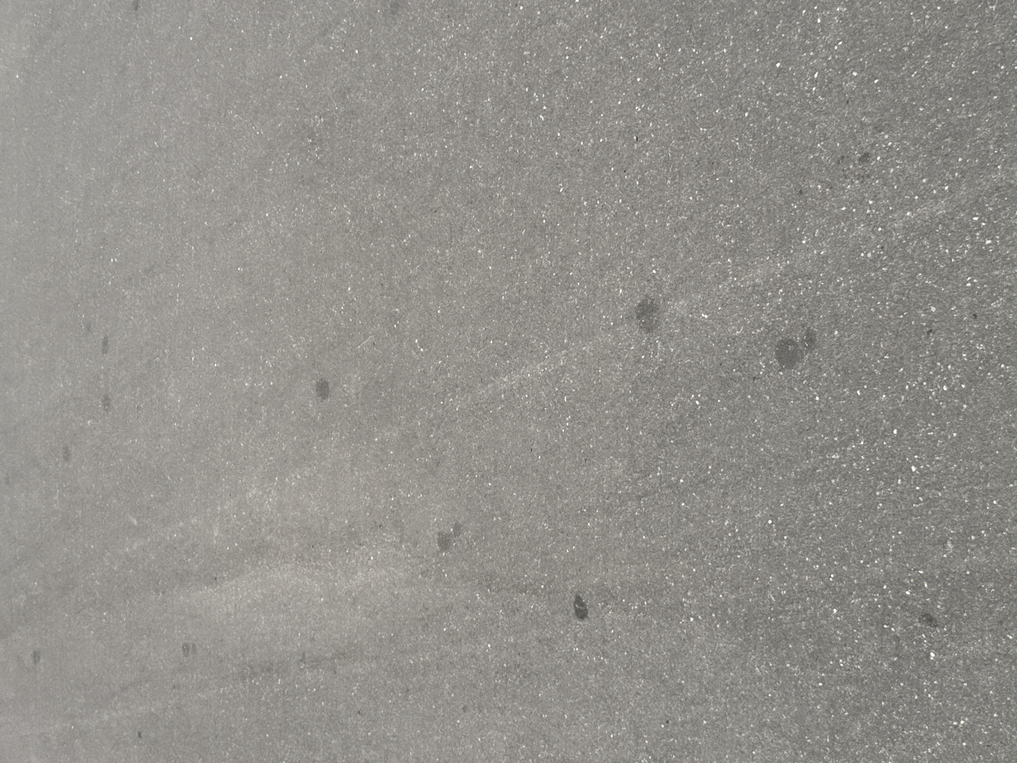 The image shows an overhead, close-up view of asphalt pavement. The asphalt is dark gray with a rough texture, speckled with small, lighter-colored aggregate. There are a few dark spots, possibly stains, scattered across the surface. No people, vehicles, or other discernible objects are present in the frame. The lighting suggests an outdoor environment. The image does not provide any contextual information that would allow for a specific location in Moscow, Russia, to be identified.
