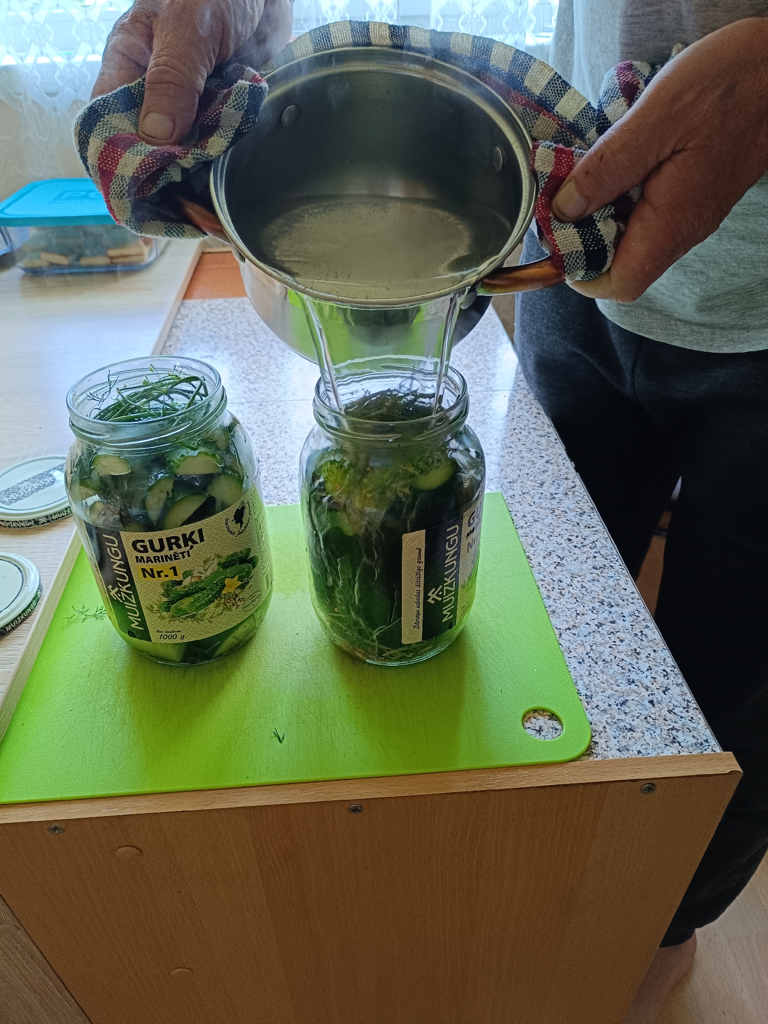 In the city of Tebras, Latvia, a person is shown in a kitchen environment pouring hot water from a stainless steel pot into a glass jar filled with pickled cucumbers and dill. The jar, labeled "GURKI MARINĒTI Nr. 1" and "MUŽKUNGU," contains sliced cucumbers and fresh dill sprigs. Another similar jar is partially visible to the left. The scene takes place on a granite countertop, with a green cutting board positioned in the foreground. Behind the jars, a light-colored window with white lace curtains is visible, and a blue plastic container with a lid rests on a wooden surface. The person's hands, wearing a patterned cloth to handle the hot pot, are the primary focus of the action. The overall setting suggests a domestic activity related to food preservation.