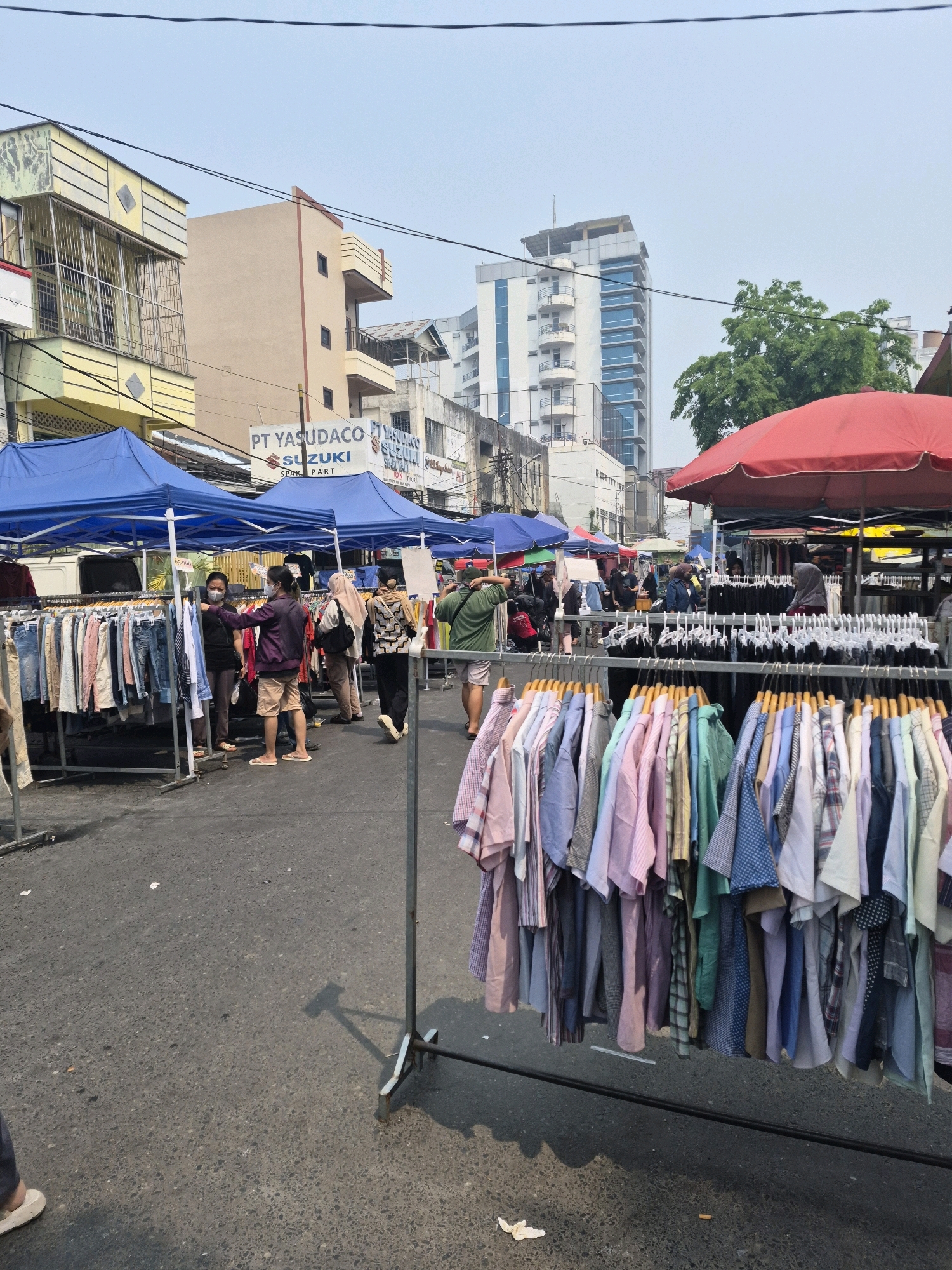 This is a ground-level, eye-level, medium shot of a street market in Banjarmasin, Indonesia. The market is set up along a street lined with multi-story buildings. In the foreground, racks of clothing are displayed for sale. The clothing consists of a variety of shirts and pants, in various colors and patterns, neatly hung on hangers. Some of the shirts are short-sleeved and appear to be casual wear, while others are long-sleeved and look more formal. In the background, several blue tents are set up, under which more clothing is displayed. People are seen browsing the stalls, with some wearing face masks. A prominent sign reads "PT YASUDACO SUZUKI SPARE PART". The overall atmosphere suggests a busy, outdoor market on a clear day.