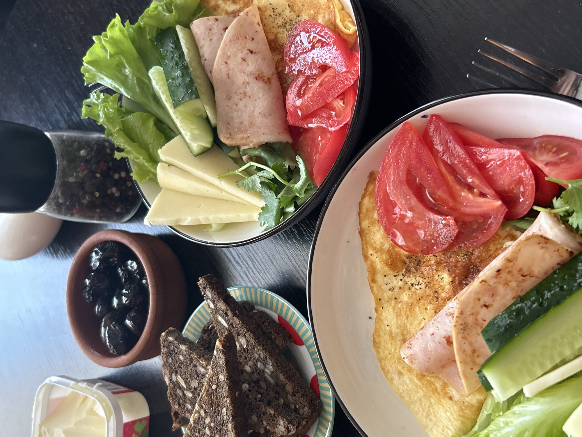 The image shows a breakfast setting. Two plates are visible, each containing a folded omelet, tomato slices, cucumber sticks, and slices of processed meat. One plate also has lettuce and cheese slices. To the side, there's a small clay pot filled with black olives, slices of dark bread on a small plate, and a container of butter. A pepper grinder and an egg are also visible on the table. The scene is shot from an overhead angle, capturing a variety of food items arranged for a meal. The location and city of Saint Petersburg, Russia are not evident from the image.