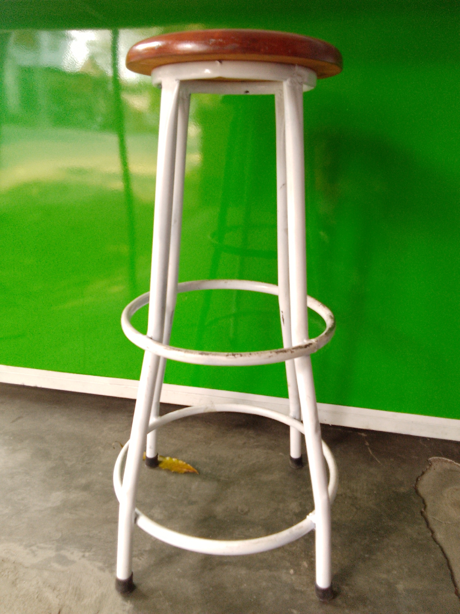 A white metal bar stool with a brown wooden seat stands on a concrete floor in front of a bright green wall. The stool has four legs connected by a circular footrest. A single yellow leaf lies on the floor near the stool's legs. The background wall appears to be reflective, showing some blurred light and shapes. The scene is likely outdoors or in a covered area due to the concrete floor and the quality of light. There are no people or animals visible. The setting does not provide specific information to pinpoint its location within the city of Trenggalek, Indonesia.