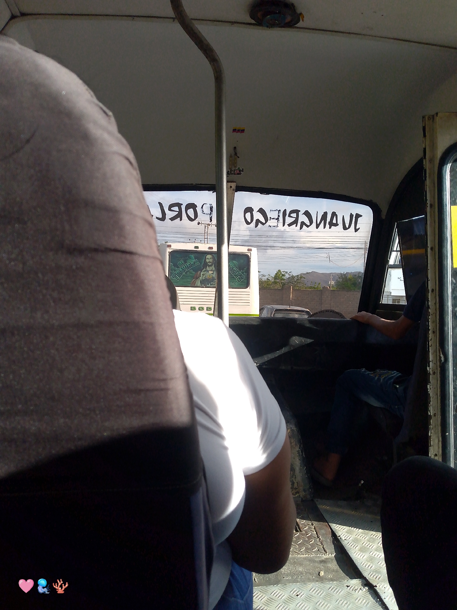 This image was taken inside a bus in Santa Ana, Venezuela. The interior of the bus is visible, with seats and windows. Outside the bus, a street scene with other vehicles and buildings can be seen. The text on the back window of the bus reads "JUANRIEGOS". A person is visible in the driver's seat. The overall lighting suggests it is daytime.