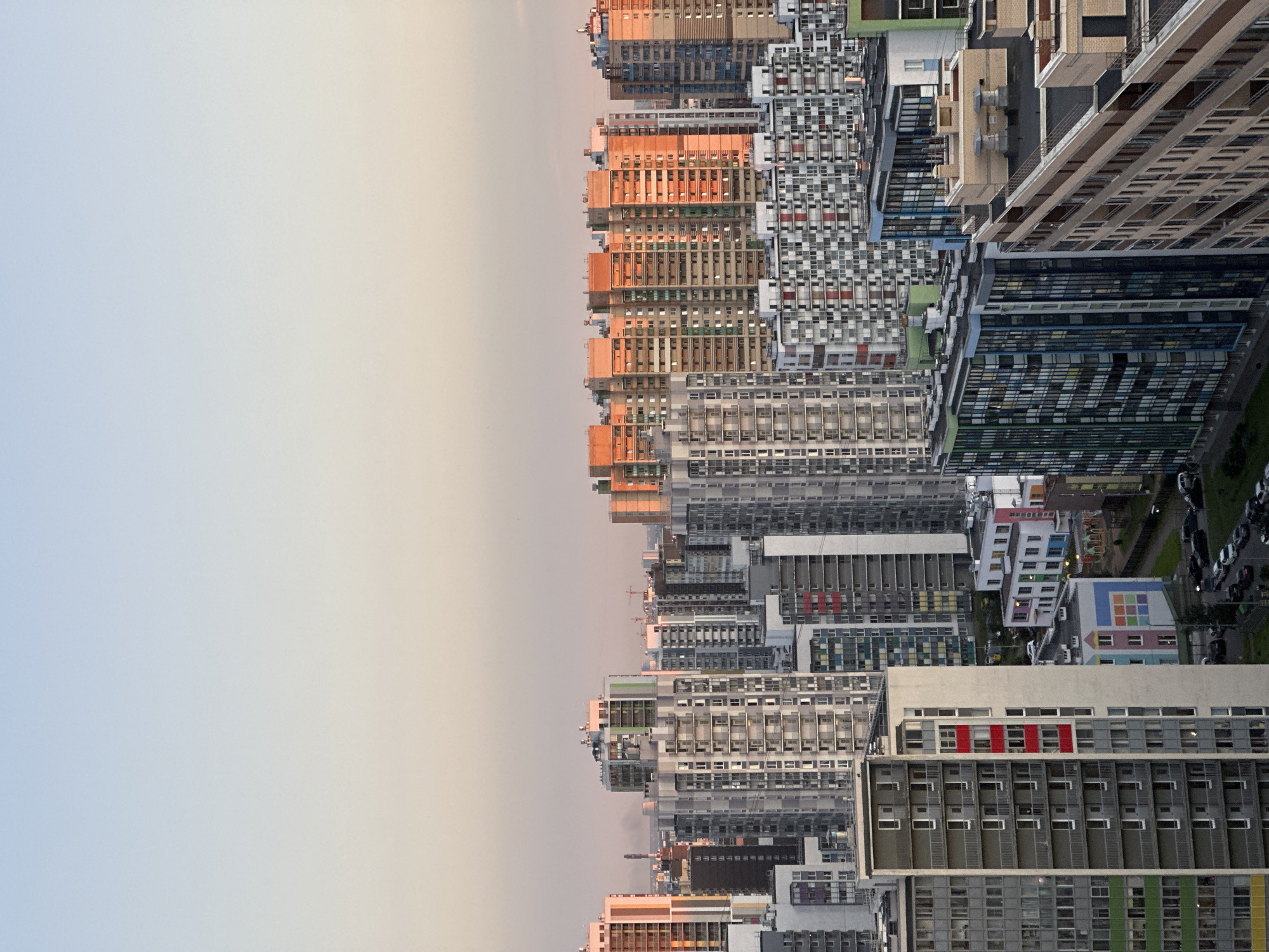 The aerial view captures a densely populated urban landscape under a pale sky, transitioning from light blue at the top to a soft pinkish hue near the horizon. A vast expanse of residential buildings, predominantly high-rise apartment blocks, dominates the scene. The buildings exhibit a variety of architectural styles and colors. Some are clad in neutral tones of gray and white, featuring numerous balconies and windows. Others are painted in warmer colors like orange and pink, illuminated by what appears to be the golden hour light. Numerous structures are interconnected or clustered closely together, creating a complex urban fabric. In the foreground, a long, narrow building with a light-colored facade and balconies with red accents stands prominently. Further back, a cluster of smaller, brightly colored buildings, possibly schools or community centers, are visible. The ground level reveals a network of roads with several cars parked or driving. The overall impression is of a modern, sprawling residential area in a Russian city.