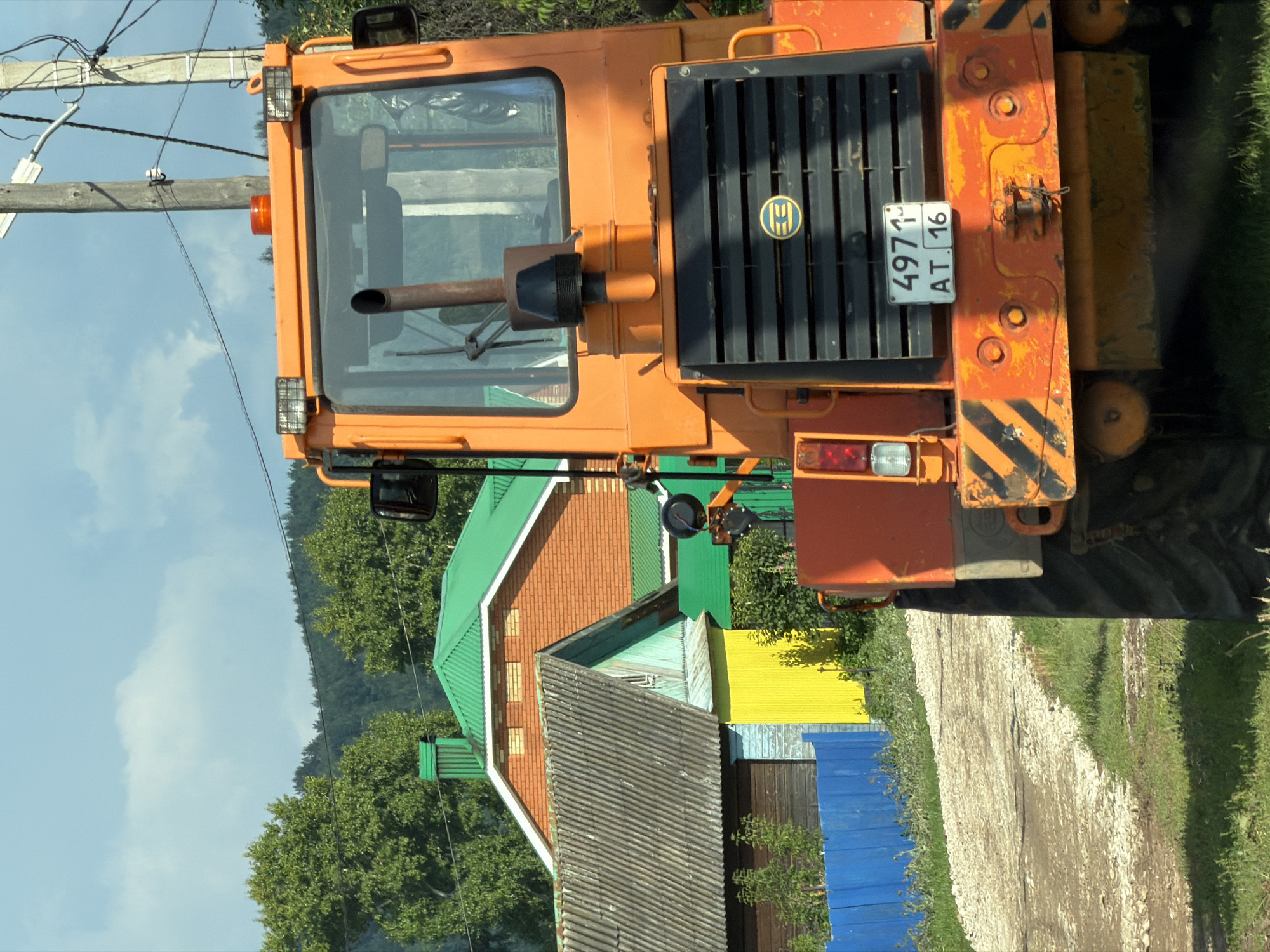 In an outdoor scene, an orange tractor is parked on a gravel road. The tractor's rear is facing the viewer. The vehicle has a black grille with a circular logo in the center, and a license plate that reads "4971 AT 16". The background includes several houses with brightly colored roofs, trees, and a cloudy sky. A wooden utility pole with wires is visible to the right of the tractor. The overall environment appears to be a rural or suburban setting.