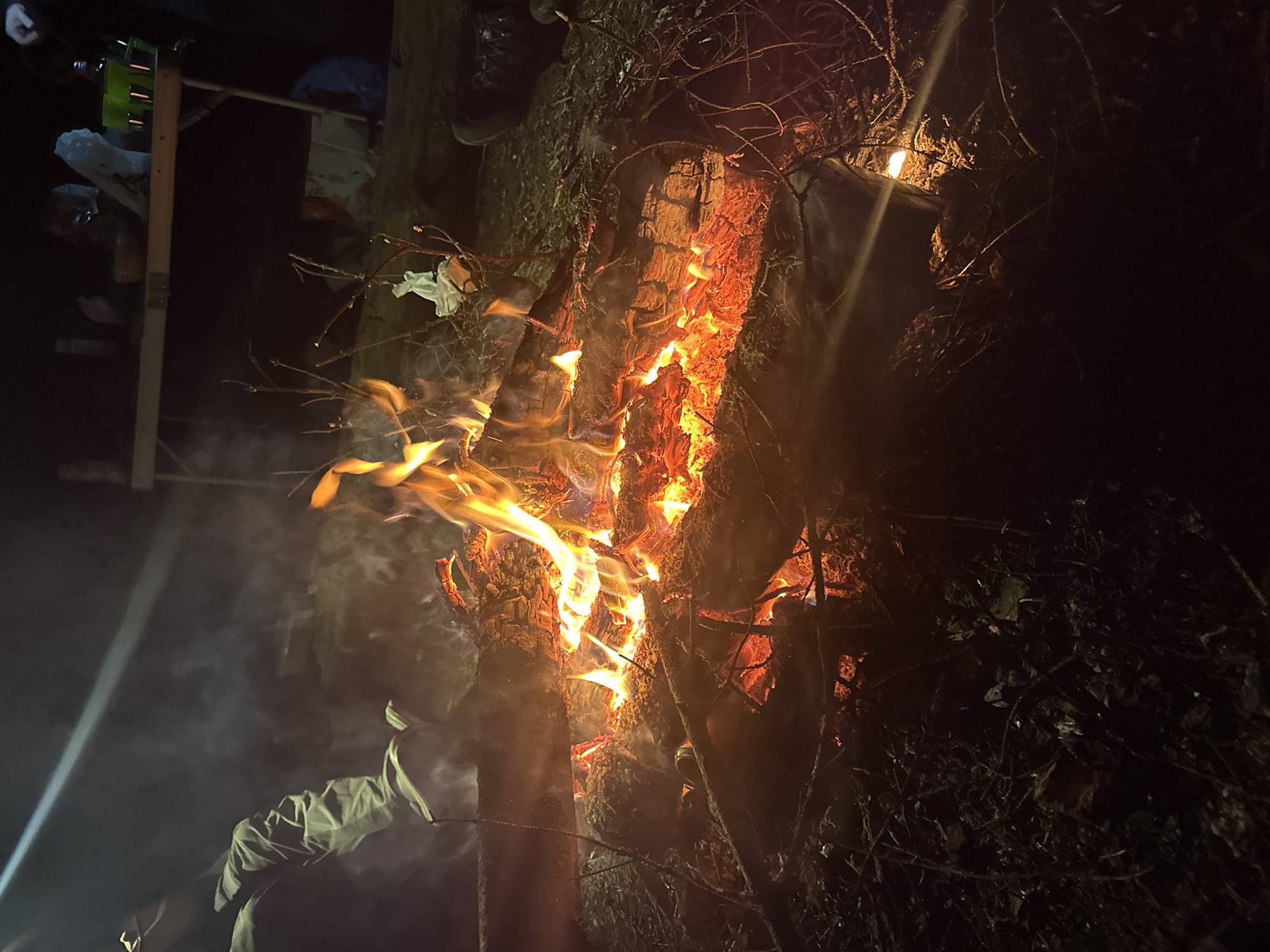 A night scene in Юзуфоўскі сельскі Савет, Belarus, shows a tree engulfed in flames. The fire is concentrated on the tree's trunk. There is smoke emanating from the base of the fire. The surrounding area is dark, with some objects partially visible in the background: a wooden structure and some unidentified items placed on top of it, as well as some darker vegetation.