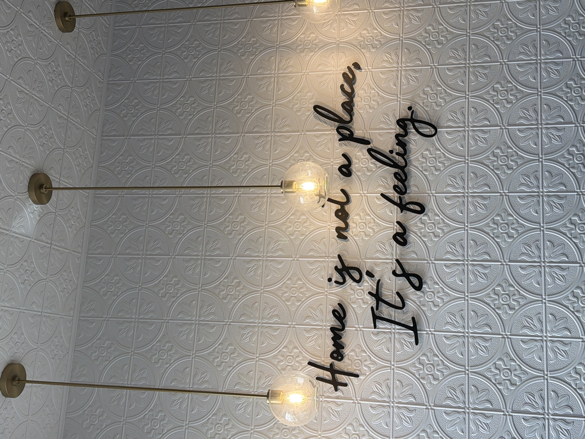 A straight-on shot shows a section of a white wall, possibly a ceiling, adorned with a repeating pattern of textured, circular floral designs arranged in a grid. The grid is formed by slightly raised lines that create squares. Three decorative, modern-style lighting fixtures are vertically aligned on the left side of the frame. Each fixture features a long, slender gold-colored rod extending from a gold circular base, with a clear glass globe enclosing a visible light bulb at the end.
Superimposed on the wall’s surface is a black, stylized script that reads, “Home is not a place, it's a feeling.” The script is oriented vertically, with each word stacked on top of the other. The text is decorative and appears to be part of an interior design element.
The lighting fixtures are placed parallel to the text, enhancing the decorative aesthetic of the wall. The overall composition focuses on the contrast between the smooth, rounded shapes of the lights and the ornate, patterned backdrop, with the inspirational text adding a layer of sentimentality. The photograph likely represents an interior shot of a retail space, cafe, or home setting.
There is no information about this image to suggest that it is located in the city of New York, United States of America.
