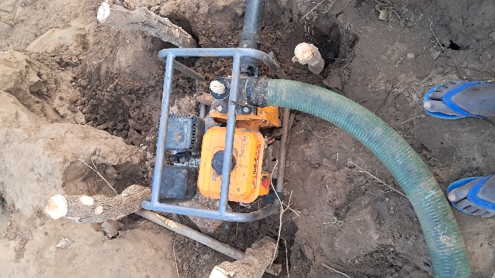 In Walu, Kenya, a scene is captured depicting an orange gasoline-powered water pump, enclosed within a grey metal frame, positioned in a depression of disturbed, dry brown earth. A dark, ribbed intake hose is connected to one side of the pump, while a lighter green, ribbed discharge hose extends from the opposite side, arcing towards the foreground. The immediate environment consists of loose soil, large clumps of earth, and several freshly cut wooden stumps. In the lower foreground, the bare feet of an individual, wearing blue flip-flops, are visible, positioned directly above the pump, indicating human presence in close proximity to the equipment.