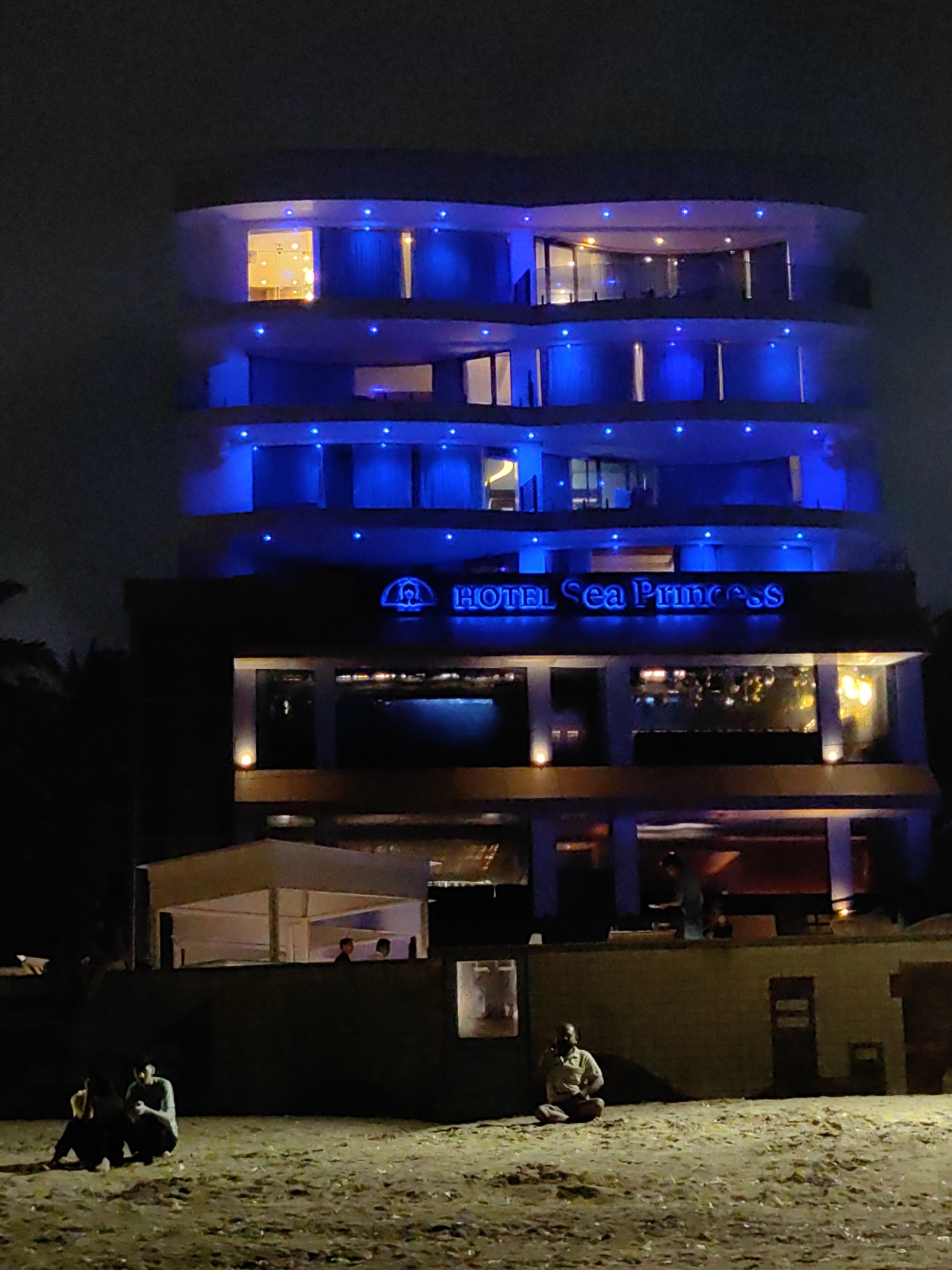 The image shows the Hotel Sea Princess in Mumbai, India, at night. The hotel is illuminated with blue lights. In the foreground, two people are sitting on the ground. A man is sitting cross-legged, holding a phone to his ear. Another group of people are visible in the background, standing near a structure. The scene appears to be on a beach, with sand in the foreground and palm trees on the left side of the image.