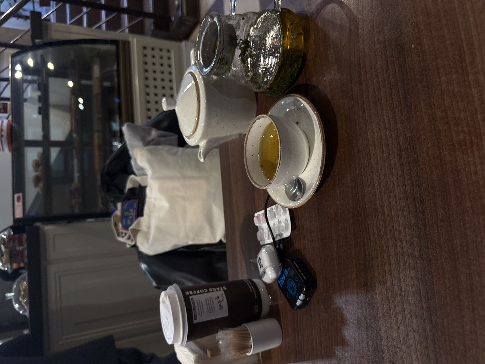 The image depicts a table in what appears to be a cafe. On the table are tea service consisting of a ceramic teapot, a glass teapot, a tea cup with a saucer, a Stars Coffee branded disposable cup with a lid and straw, a small cleaning brush, and a small black object which appears to be an electronic device with a screen. Additionally, there are ear buds and a charging cord. In the background, there is a glass display case with food items inside, and architectural details suggesting an indoor location. Based on the brand of coffee cup, the location is likely in the city of Moscow, Russia.