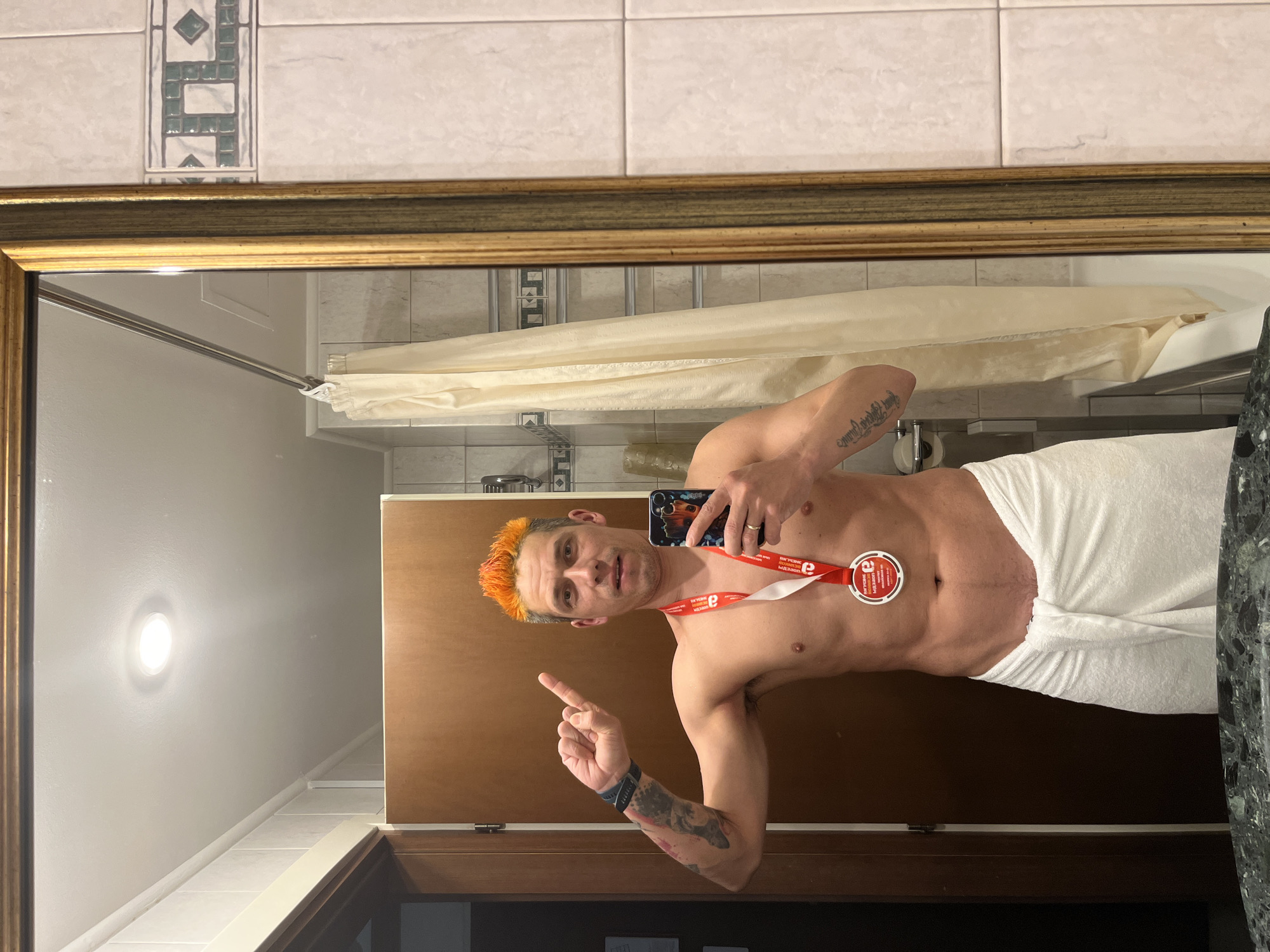 A man with orange hair takes a selfie in a bathroom mirror. He wears a white towel around his waist and a medal around his neck. His torso is bare and muscular, revealing abs. He points with one finger. The bathroom has tiled walls and a shower curtain.