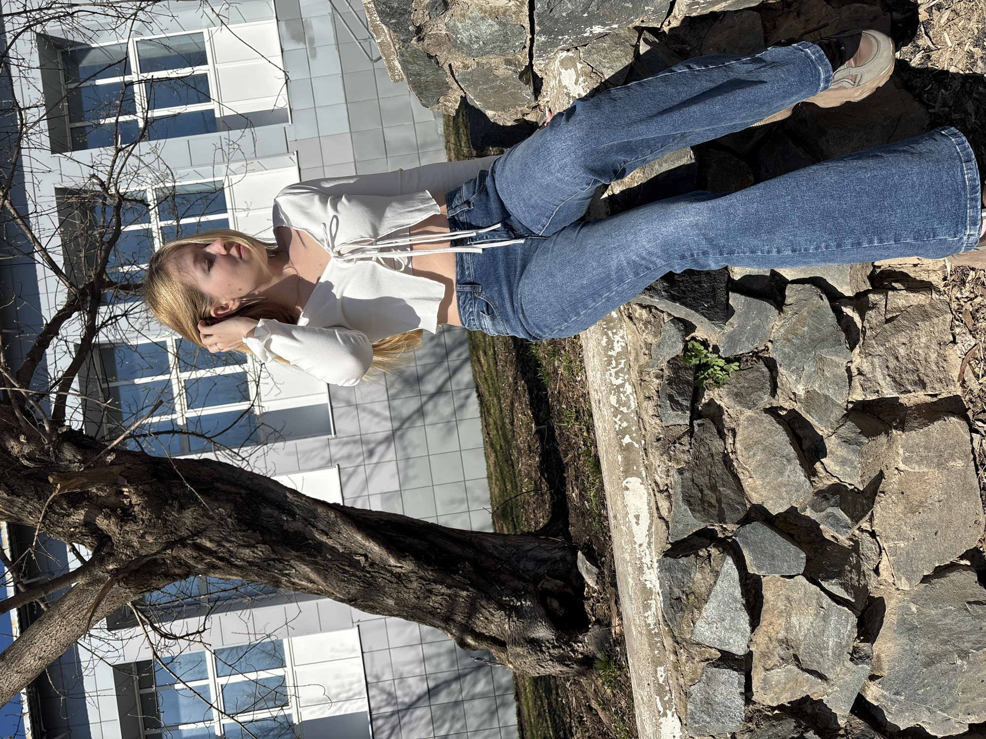 This image captures a young woman posing outdoors on a bright, sunny day, likely in Chernushka, Russia, given the context. The main subject is a fair-skinned woman with long, straight blonde hair, dressed in a white, long-sleeved ribbed crop top with front ties, paired with blue jeans and light-colored chunky sneakers. She is seen looking upward and slightly to her right, with one hand gently touching her hair, conveying a serene or contemplative mood.
The setting appears to be a landscaped area, possibly a park or an urban green space, adjacent to a modern building. The woman is positioned next to a rustic wall or embankment constructed from various sizes of dark-grey rocks. To her left, a large, leafless tree with gnarled, dark branches adds a natural element to the scene. In the background, a contemporary building with numerous large windows reflects the clear blue sky, suggesting a bright and open environment. The ground beneath and around the tree consists of dry earth with sparse patches of green vegetation.
The distinct shadows and strong sunlight indicate that the photograph was taken during midday or early afternoon under clear weather conditions. There is no visible text present in the image.