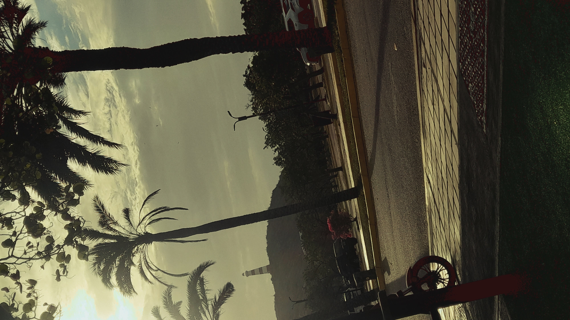 The image captures a scenic view in Pedro González, Venezuela, under a cloudy sky. In the foreground, a portion of a red scooter is visible on a sidewalk paved with light-colored rectangular tiles, casting long shadows. Adjacent to the sidewalk is a road with a yellow border, and beyond that, a grassy area. In the midground, several palm trees dominate the scene, with one particularly tall, dark palm trunk on the right. Behind the trees, a hill is visible, topped by a striped lighthouse. The landscape features a line of trees and some parked vehicles, including a dark SUV. The overall impression is that of a coastal or park-like setting in a tropical environment.