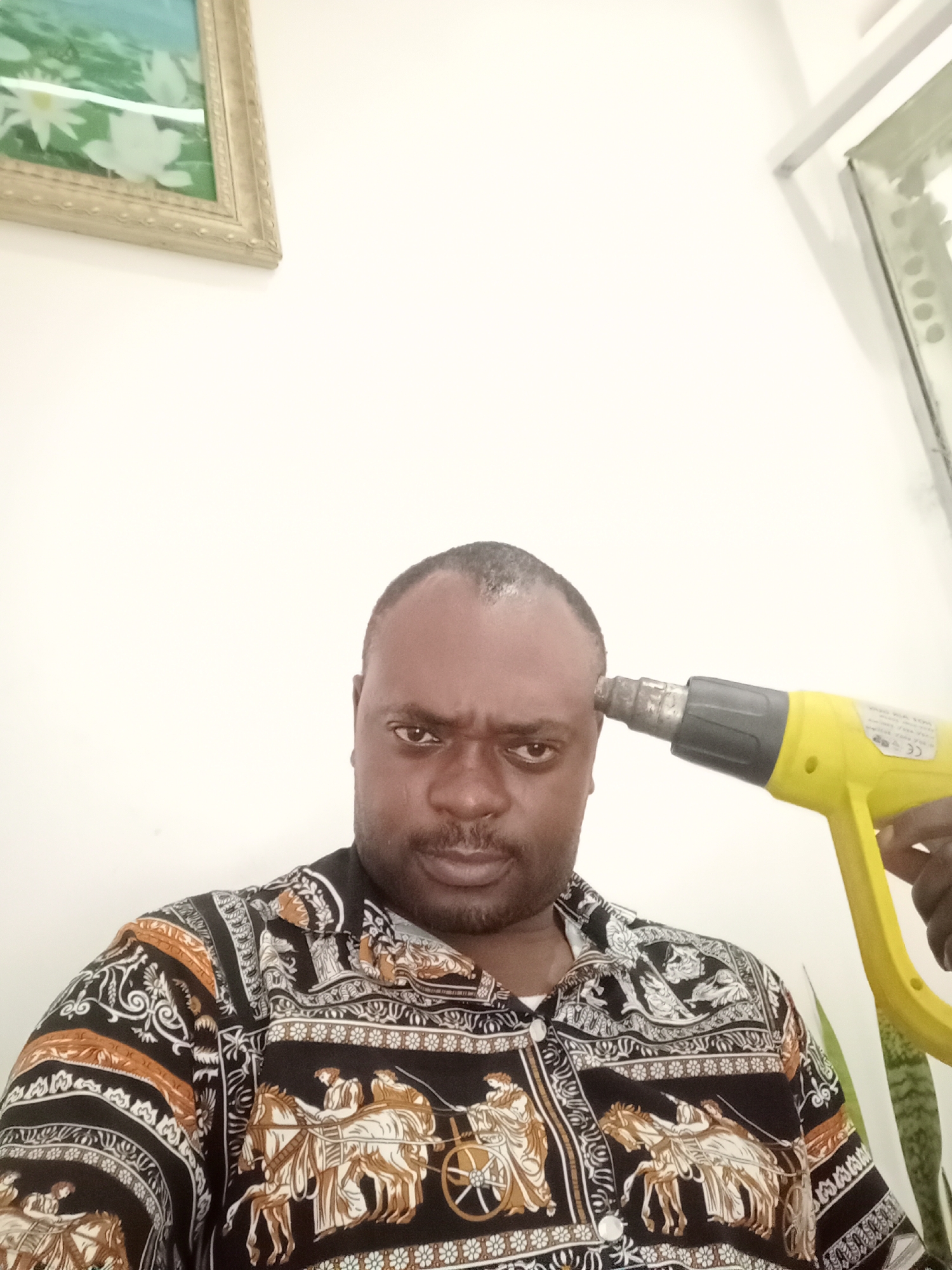 A man is holding a yellow heat gun to his forehead. He is wearing a black and orange patterned shirt. There is a picture frame on the wall behind him. The location is Gwarinpa, Nigeria.