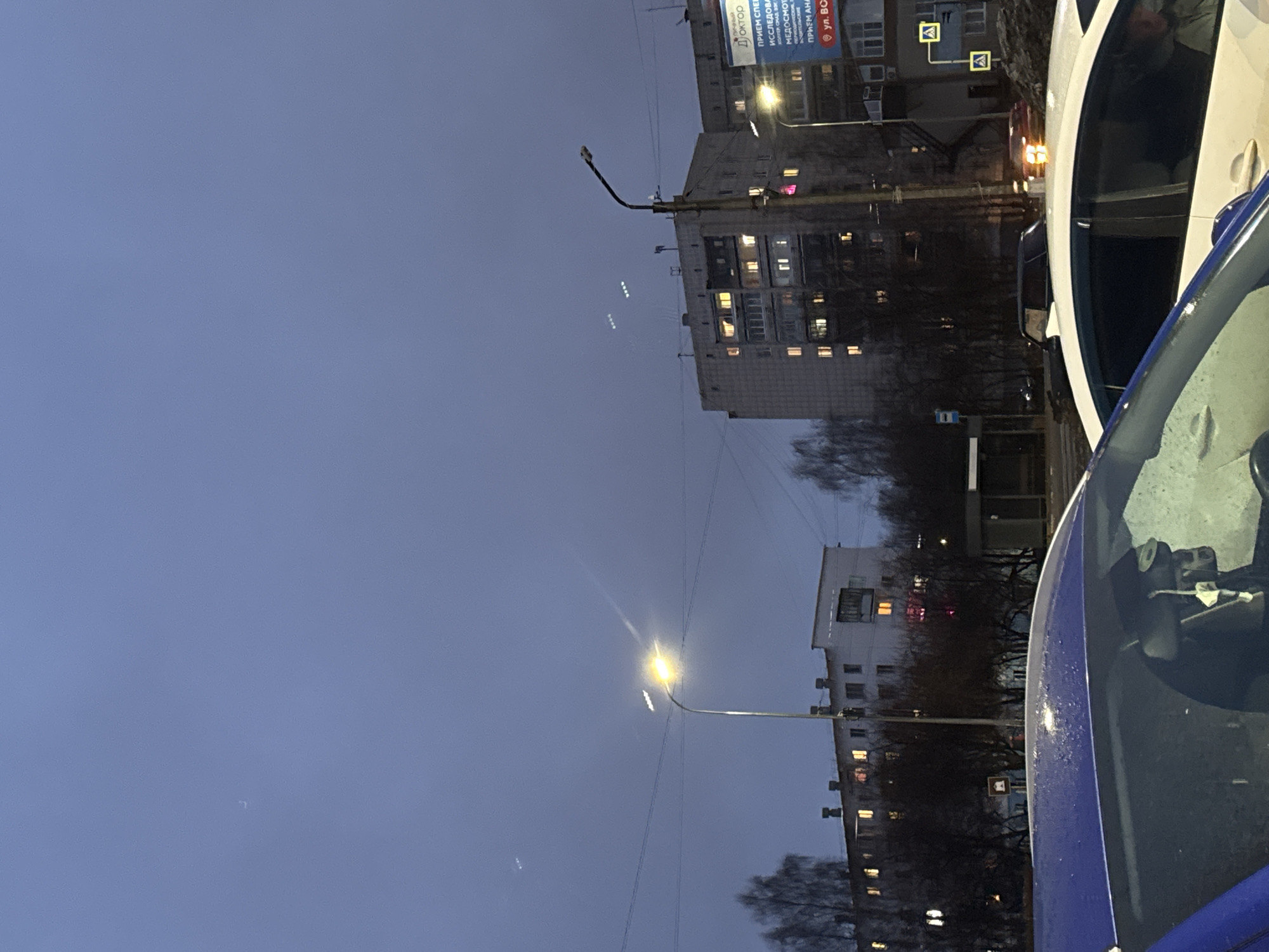 The image shows a street scene in Chaikovsky, Russia, at dusk. Two cars, one white and one blue, are parked on the side of the road. Several apartment buildings are visible in the background, with lights on in some windows. Streetlights illuminate the area. Bare trees line the street.