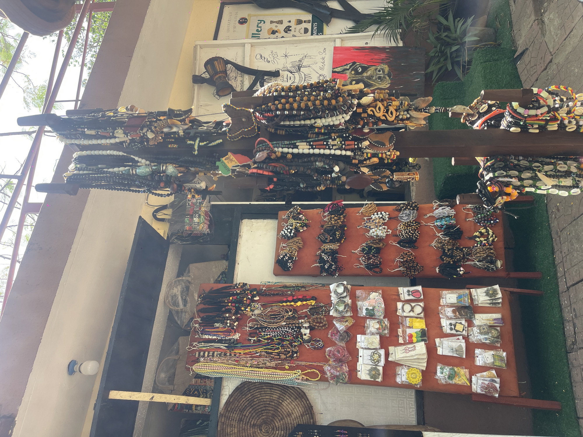 The image depicts an outdoor market stall displaying a variety of jewelry and crafts. The stall is heavily laden with necklaces and bracelets, some hanging from wooden supports and others arranged on tables. The jewelry appears to be made of beads, shells, and other natural materials. There are also packaged items on the tables, possibly earrings or small trinkets. The stall is set up in an outdoor area, possibly a street or plaza, with some greenery visible in the background. The location appears to be in a city.