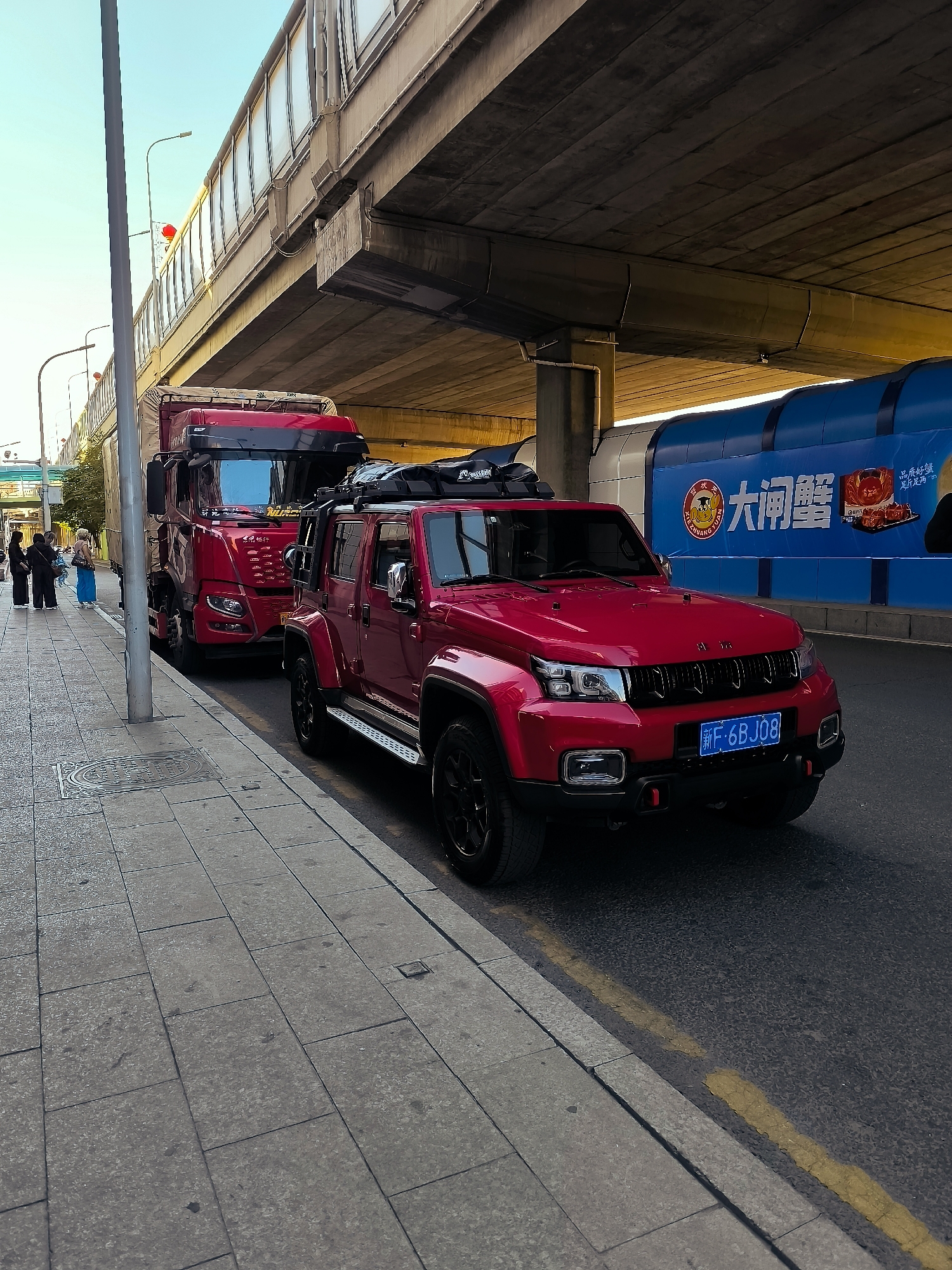 The image captures a street scene in Ürümqi, China, with a prominent overpass casting shadows. In the foreground, a bright red Jeep is parked on the right side of the road. It has a roof rack with luggage. To the left of the Jeep, a red truck is also parked. On the sidewalk to the far left, three individuals, two women and one person in a blue skirt, are standing. Behind the vehicles, a large blue billboard with Chinese text and an image of crabs is visible. The sky is a clear light blue. The overall lighting suggests it is daytime, likely late afternoon due to the long shadows. The road surface is asphalt with a yellow line marking the lane. The sidewalk is paved with large square tiles.