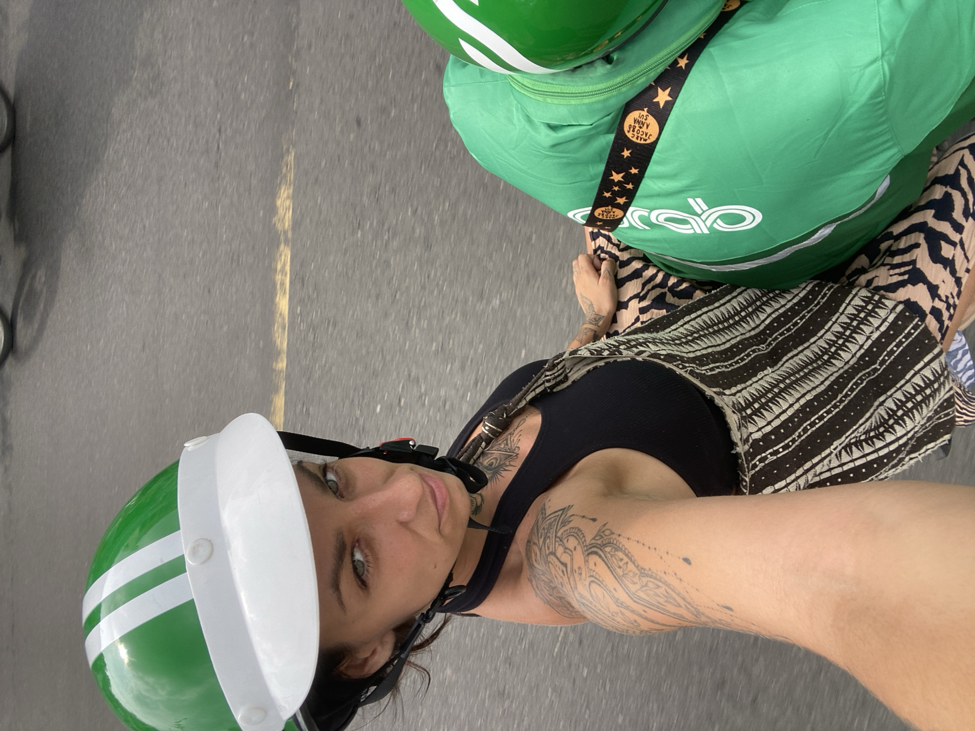 A woman wearing a green and white helmet and a black tank top is seated on a motorbike in Phu Thuy Ward, Vietnam. She has tattoos on her arm and chest. The motorbike is equipped with a large green bag, also featuring white branding and a black strap with stars. The road surface is asphalt with a yellow line. The image is taken from a first-person perspective, suggesting the woman is taking a selfie while on the moving vehicle.