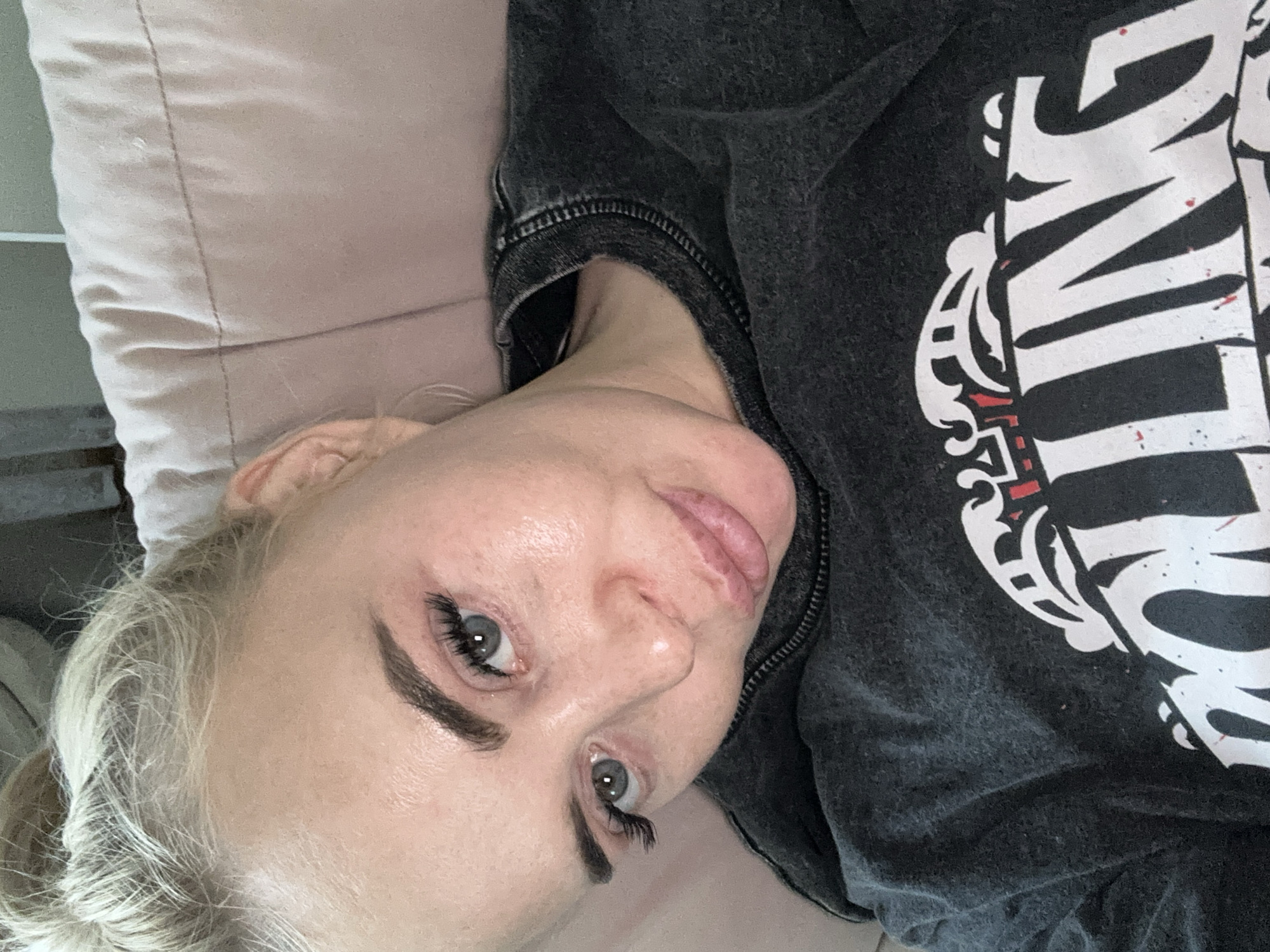 A blonde-haired person is lying down, possibly on a light pink pillow. The individual is wearing a dark gray, washed-out t-shirt with a large white graphic design on the front. The person's face is visible, with prominent eyelashes and eyebrows. The location is unspecified and not discernible from the image.