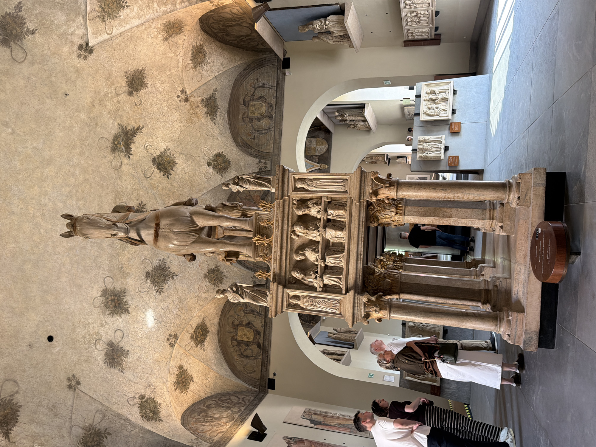The image shows a statue of a rider on horseback inside a museum. The museum has a painted ceiling with decorative floral patterns and coat of arms, with other sculpted objects and historical displays visible in the background. Several people are present, seemingly tourists or museum visitors, observing the statue and exhibits. The statue itself is intricately carved with scenes and figures, resting on a pedestal supported by columns. Based on the architecture and artifacts displayed, the location is likely the Museo di Arte Antica in Milan, Italy, within the Sforza Castle complex.