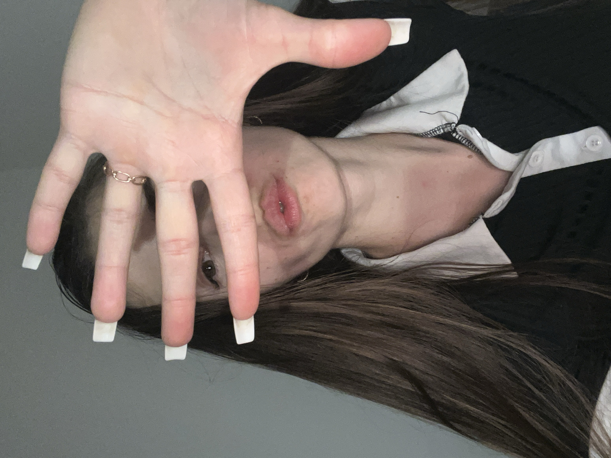 An unidentified female with long, dark brown hair is captured in a medium shot, posing with her right hand held up to obscure her face. She wears a black garment with a white collar and buttons. Her fingers are adorned with long, white, square-shaped acrylic nails and a delicate rose-gold ring with an oval link design is visible on her ring finger. Her lips are slightly parted and painted with a pink lipstick. The background consists of a plain, gray wall, suggesting an indoor setting. The lighting is dim, casting soft shadows.