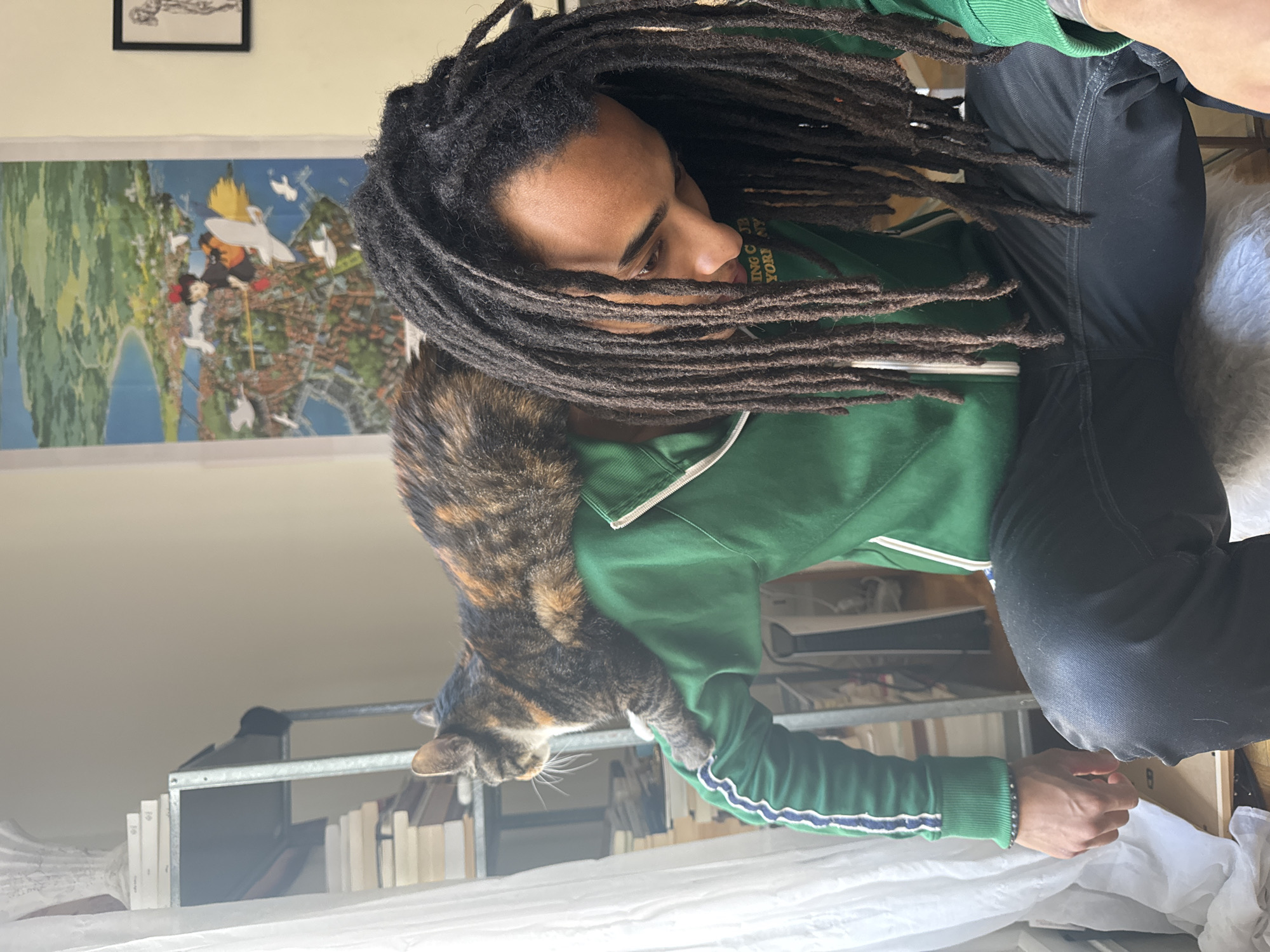A medium, indoor shot depicts a man with long dreadlocks wearing a green zip-up jacket and black pants, seated with a calico cat draped across his left arm and shoulder. The man looks down, his face partially obscured by his hair.
Behind him, partially visible, is a framed artwork depicting an illustration of a city, resembling Paris, featuring landmarks and stylized elements, hung on a white wall. Above this, another framed picture is barely visible. To the left, a metal shelving unit stacked with books is present. The lower portion of the image shows a white cloth or sheet draped across objects, possibly furniture or storage. The lighting appears to be natural, with bright daylight entering from the left side of the frame. The cat's presence suggests a domestic setting, possibly an apartment.