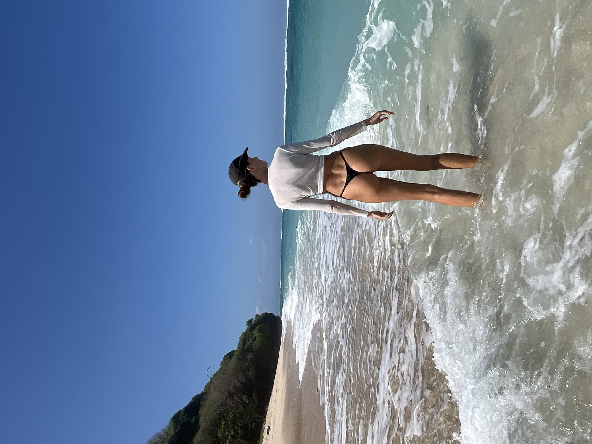 A woman stands in shallow, clear ocean water on a sandy beach. She is facing away from the camera, looking out at the turquoise ocean. She is wearing a white, long-sleeved, cropped shirt and a black thong. A dark baseball cap is on her head. The beach is backed by a lush, green cliff face. The sky is a clear, bright blue. The scene appears to be the southern coast of Bali, Indonesia, a popular tourist destination known for its beaches.