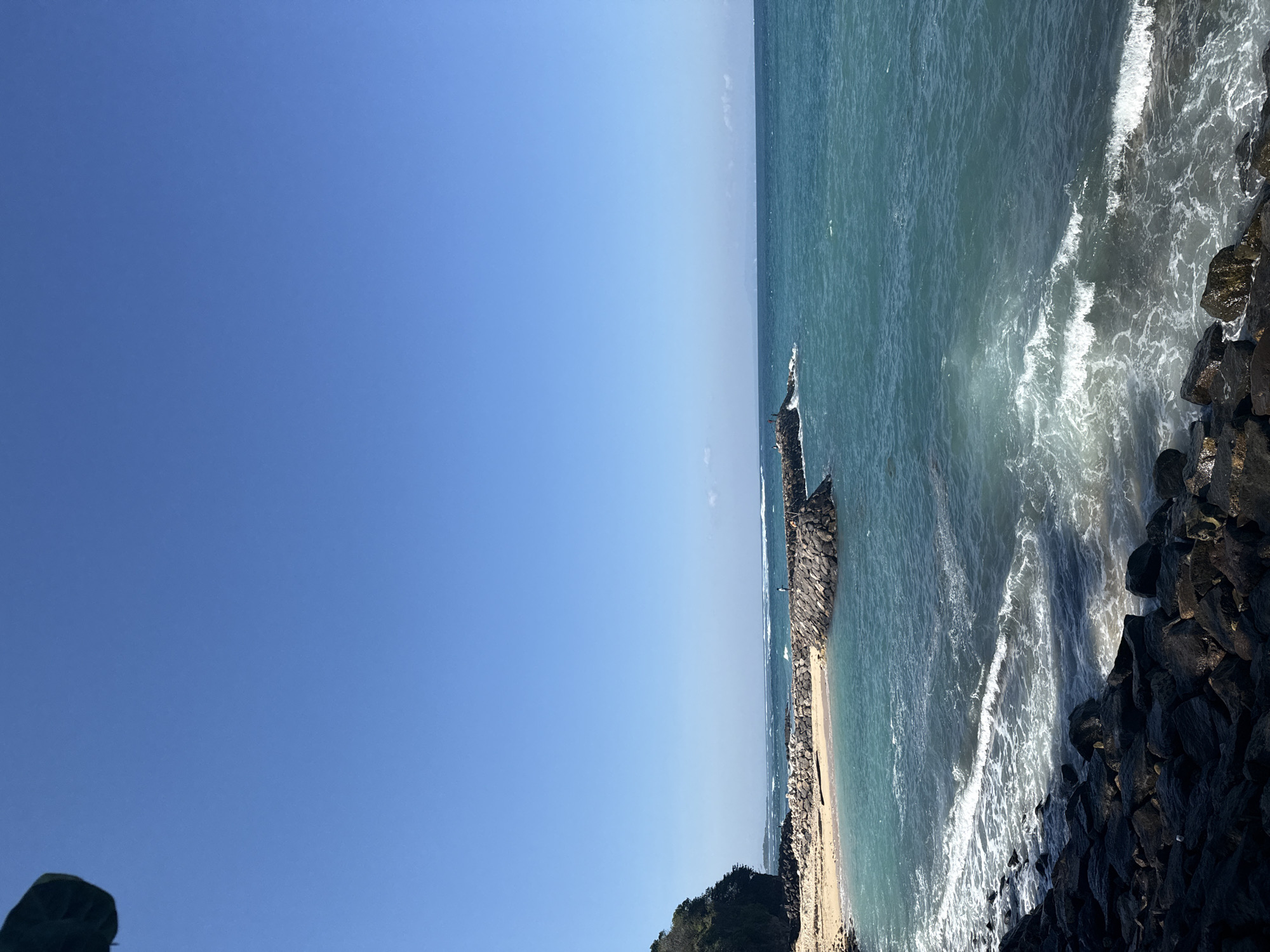 This image displays a coastal landscape in Kuta Selatan, Indonesia, under a clear, cloudless blue sky. The foreground is composed of dark, irregularly shaped rock formations along the immediate shoreline, where white-crested waves are breaking. Above these rocks, a section of light brown sand forms a beach, bordered on the far left by a verdant, treed cliff or hill. Extending from the sandy area into the teal-colored ocean are multiple man-made rock jetties, constructed from large, grey boulders. One prominent jetty arcs into the water, and a single human figure is discernible standing on its furthest visible point. Further to the right, another smaller rock formation or jetty hosts approximately two to three indistinct human figures. The ocean exhibits various shades of blue and turquoise, with white-capped waves breaking both near the shore and further out. A subtle, hazy line on the distant horizon suggests either a very distant landmass or atmospheric conditions. The upper two-thirds of the image consist entirely of a bright, uniform blue sky. A dark, out-of-focus, curvilinear object, possibly an umbrella or foliage, intrudes into the extreme upper-left corner of the frame.