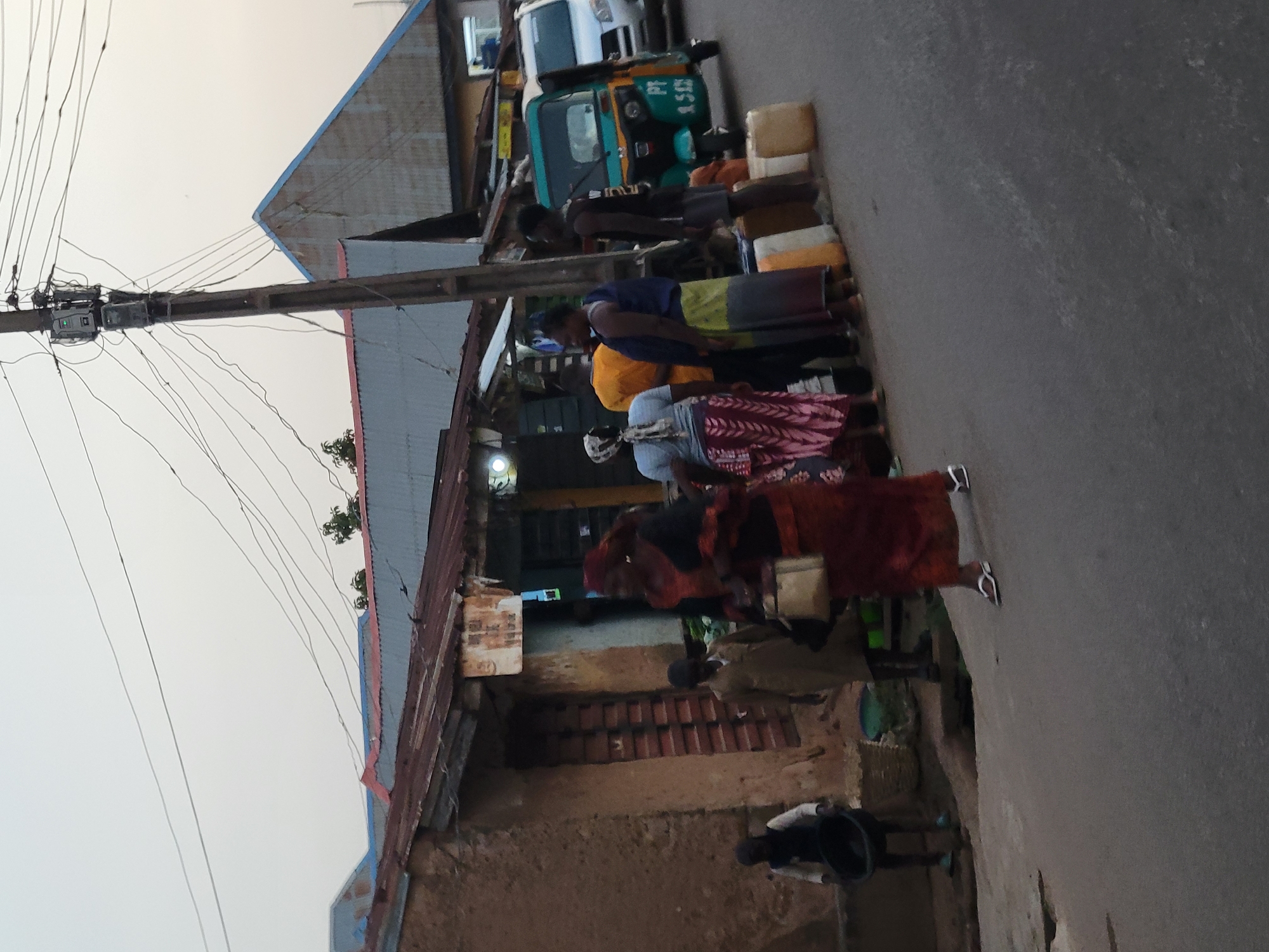 An outdoor street scene in Jos, Nigeria, depicts daily life along a paved road, likely in the early morning or late afternoon given the soft, diffused light and hazy sky.
Several people are present along the roadside. A group of adults stands near a line of yellow jerrycans. One person wears a blue shirt and a yellow patterned skirt, another a white top and a distinctively patterned skirt, and a third is dressed in a red or maroon garment. A person in a dark top and shorts is positioned near the jerrycans, possibly attending to them. Further down the road, another individual is partially visible inside a parked vehicle.
Prominent objects include numerous yellow jerrycans, suggesting a point for water or fuel collection/distribution. A green and white three-wheeled auto-rickshaw (commonly known as a Keke Napep in Nigeria) is parked, with part of a white van or truck visible behind it. A tall utility pole stands on the left, heavily strung with numerous electrical wires and what appear to be connection boxes or small transformers. Buildings with corrugated metal roofs and textured, rustic walls line the street. On the ground, near the buildings, a black bucket and a woven basket are visible.
The scene captures a typical urban or semi-urban activity in Nigeria, likely involving the transport or sale of essential commodities like water or fuel, indicated by the jerrycans. On the side of the three-wheeled auto-rickshaw, the text "PT 9536" is visible, likely an identification or license number for the vehicle.