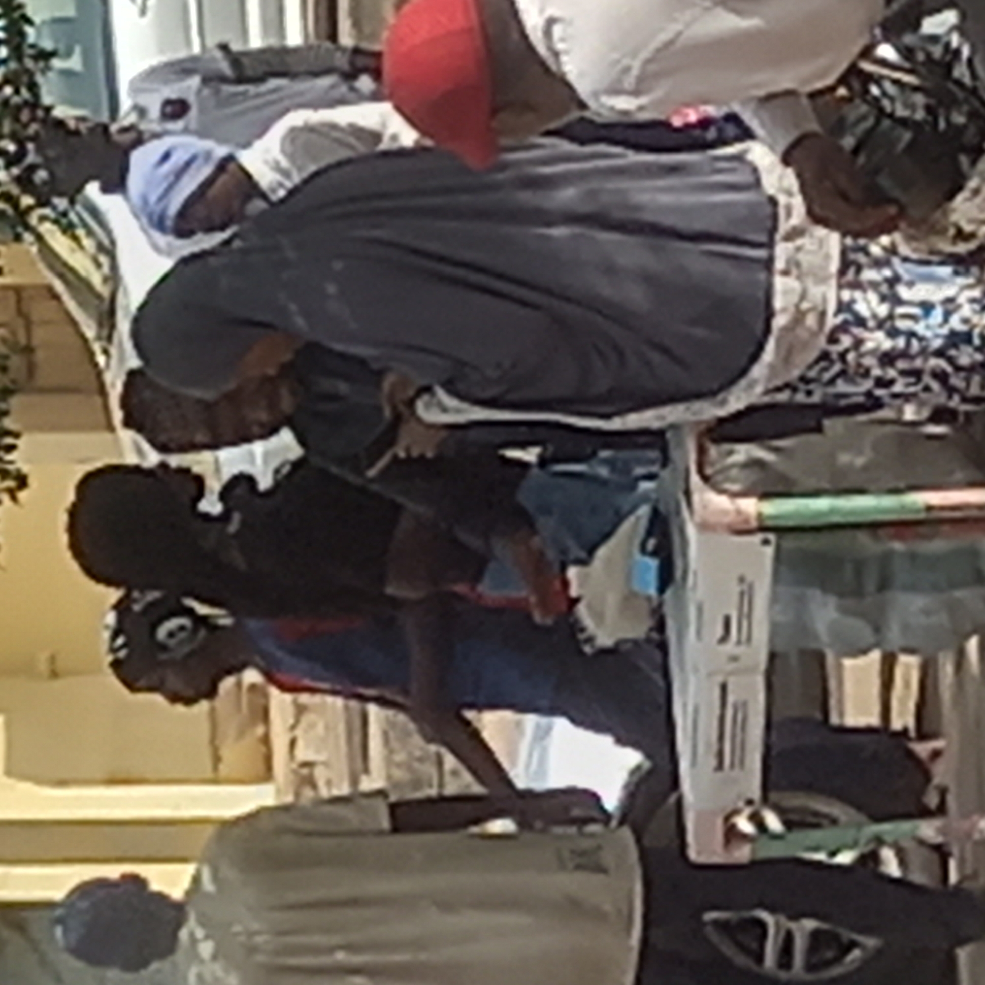 In the city of Sardauna, Nigeria, a group of people are gathered. One person wears a red cap. A woman in a dark gray garment appears to be focused on something. Another individual wears a blue cap. The group appears to be around a table or surface with what looks like some goods displayed. There is also what may be a vehicle and building in the background.
