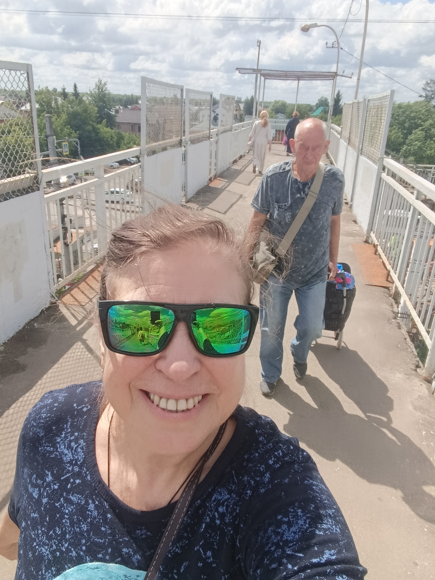 An outdoor, eye-level, medium shot captures a woman in the foreground taking a selfie on a pedestrian overpass. She has brown hair and wears large, dark sunglasses with green reflective lenses and a dark blue t-shirt with a light blue speckled pattern. Behind her, a man walks away from the camera, wearing a blue t-shirt, blue jeans, and a crossbody bag. He is pulling a black wheeled shopping cart filled with items. Further down the overpass, two women walk towards the camera; one in a light-colored dress and the other in a dark outfit. The overpass has white railings and metal mesh fencing. The background shows a cityscape with buildings and lush green trees under a cloudy sky. The scene is indicative of a sunny day. The image was taken in Orekhovo-Zuyevo, Russia.