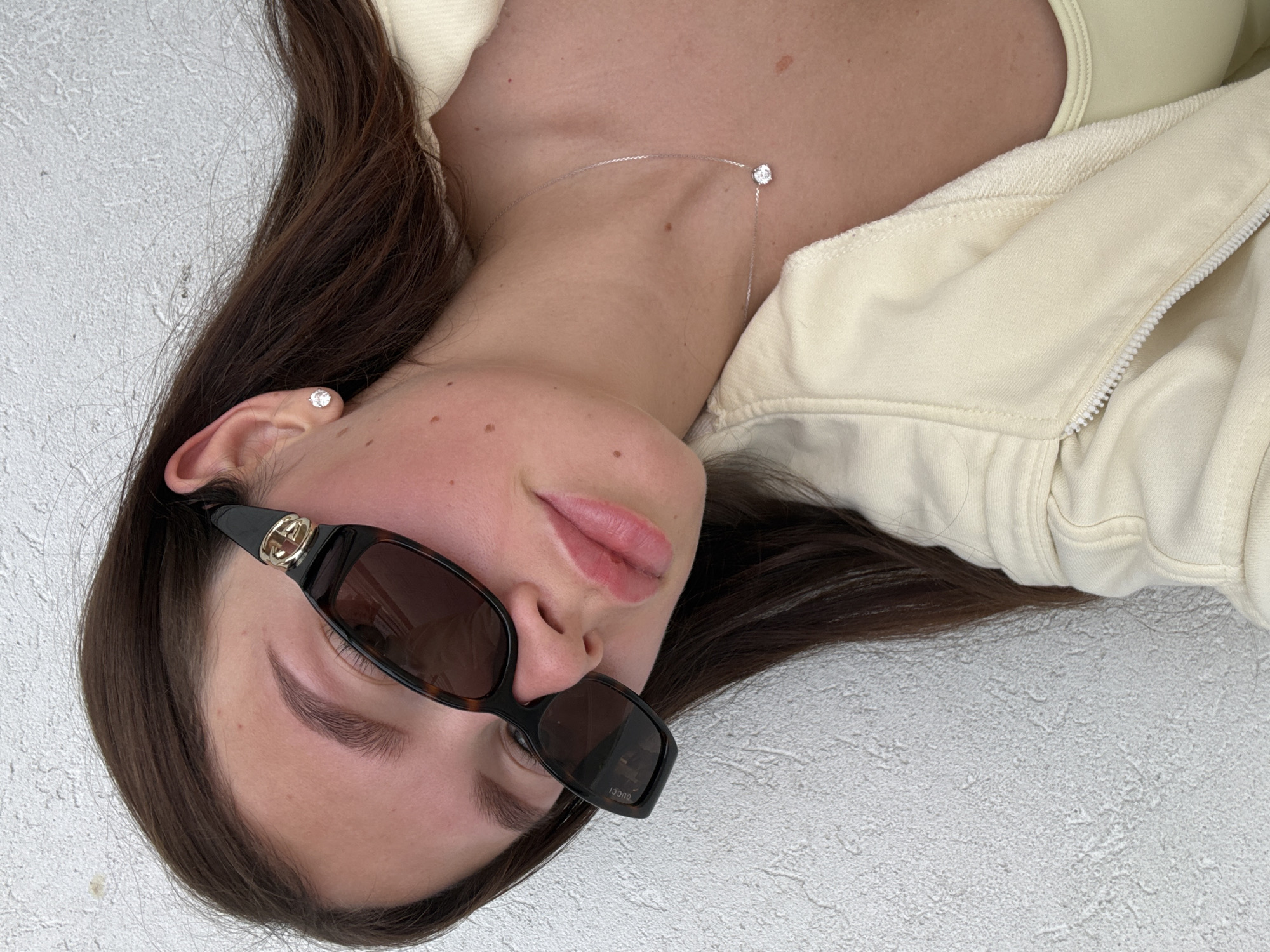 A young woman is lying on a light-colored surface, possibly concrete. She has long, dark brown hair and is wearing sunglasses with a gold-colored logo on the side. She is wearing a light yellow jacket with a zipper and a light yellow top underneath. She is also wearing a diamond stud earring and a delicate necklace with a single diamond pendant. The location appears to be outdoors, possibly in Tel-Aviv, Israel, but there are no specific visual cues in the image to confirm this beyond doubt.