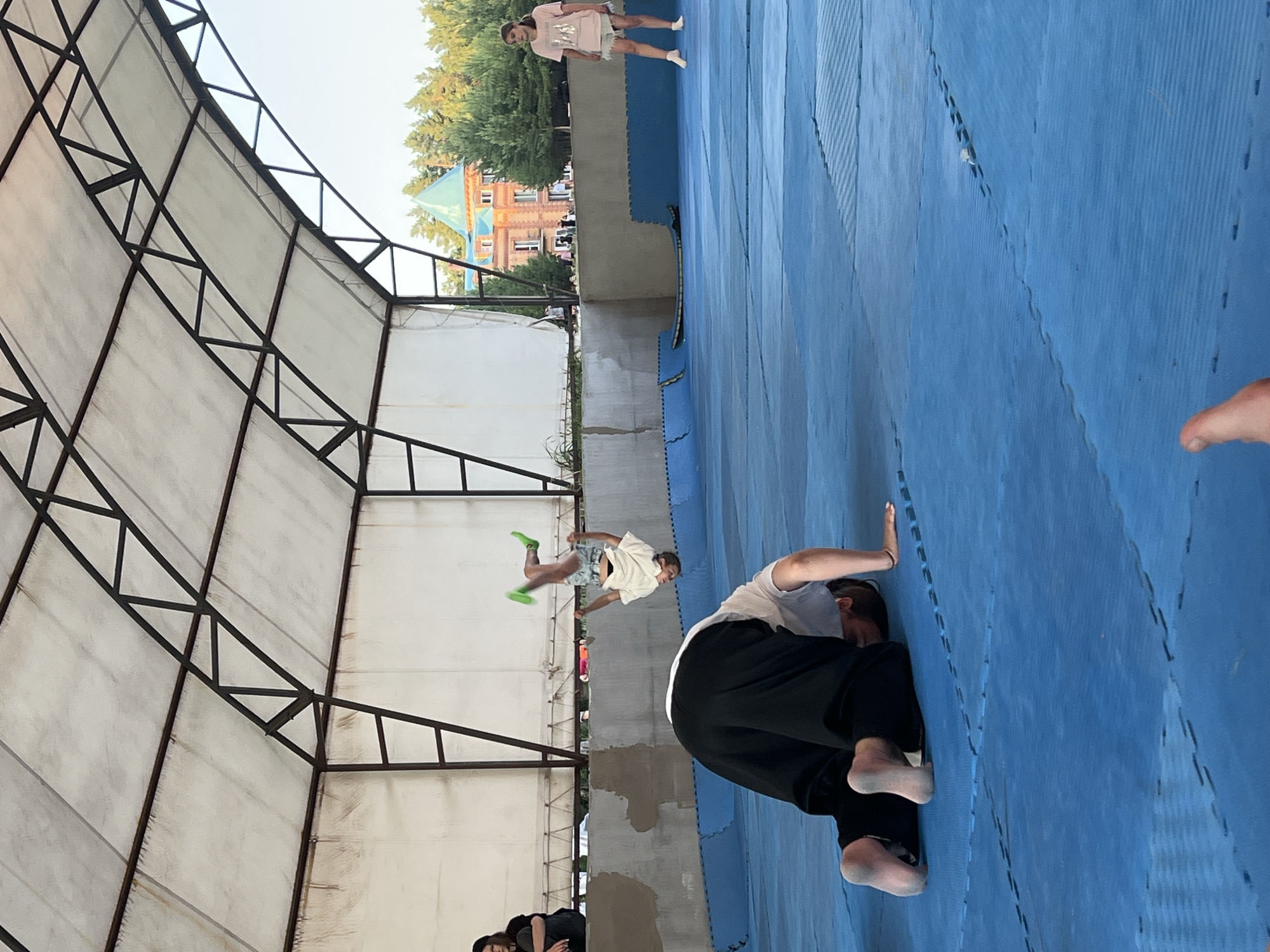The image captures an outdoor training session, likely a martial arts or parkour practice, taking place under a simple open-sided structure with a metal frame and translucent roof. The ground is covered with blue interlocking mats, commonly used for such activities.
Several individuals are present. In the mid-ground, a person is mid-air, performing a jump or flip, dressed in a white t-shirt and patterned shorts with bright green socks. Another person is on the blue mats in the foreground, in a crouched or rolling position, wearing a white t-shirt and black pants. Further in the background, another individual stands, wearing a pink t-shirt and shorts. A person's hand and arm are visible in the far right foreground, suggesting another participant or observer.
The scene appears to be set during daylight, with the sun casting shadows, indicating it's likely daytime, possibly late afternoon given the angle of the light. The background shows trees and a building with a distinctive green roof and ornate brickwork, which is identified as being in Krasnoturansk, Russia. The overall atmosphere seems active and focused, with the participants engaged in physical training. There is no visible text in the image.
