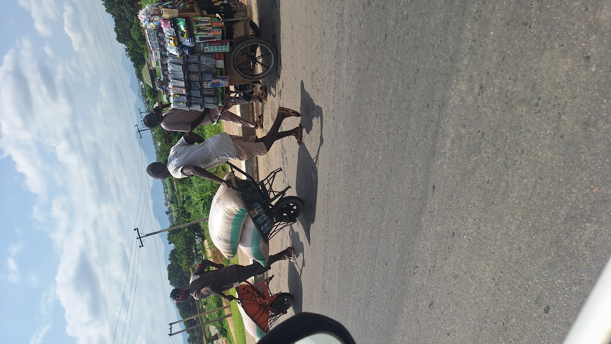 A sunny day in Jalingo, Nigeria, with a clear blue sky dotted with white clouds. In the foreground, a paved road is visible, with two men pushing wheelbarrows filled with large sacks. The man on the left wears a dark shirt and headscarf, while the man on the right wears a white t-shirt and khaki shorts. Behind them, another man in a light brown shirt stands near a cart laden with various goods, including phone accessories and other items displayed on shelves. The cart has a large spoked wheel. In the background, lush green vegetation lines the road, and distant mountains can be seen under the sky. Utility poles with wires are also present. The scene depicts everyday life and commerce on a roadside in the city.