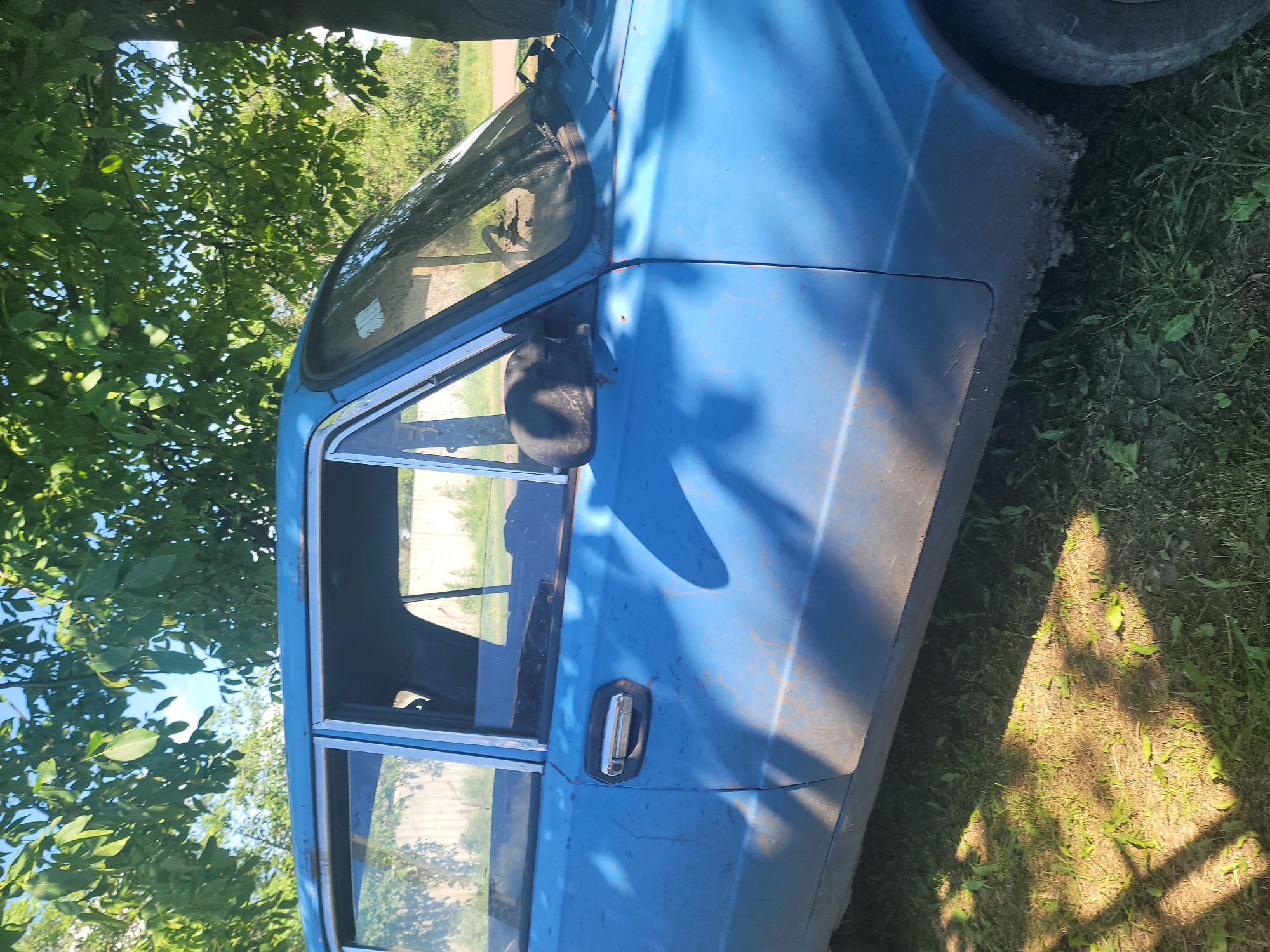 The image captures the side view of a blue, older model sedan parked on grassy ground under the dappled shade of a tree. The car appears to be in a state of disrepair, with visible rust and dirt. The background shows a white fence and lush green trees, suggesting a rural or semi-rural setting. The sky is clear blue, indicating a sunny day. The overall scene is static and devoid of any human activity. It's likely located in Vovkivka, Ukraine, given the context.
