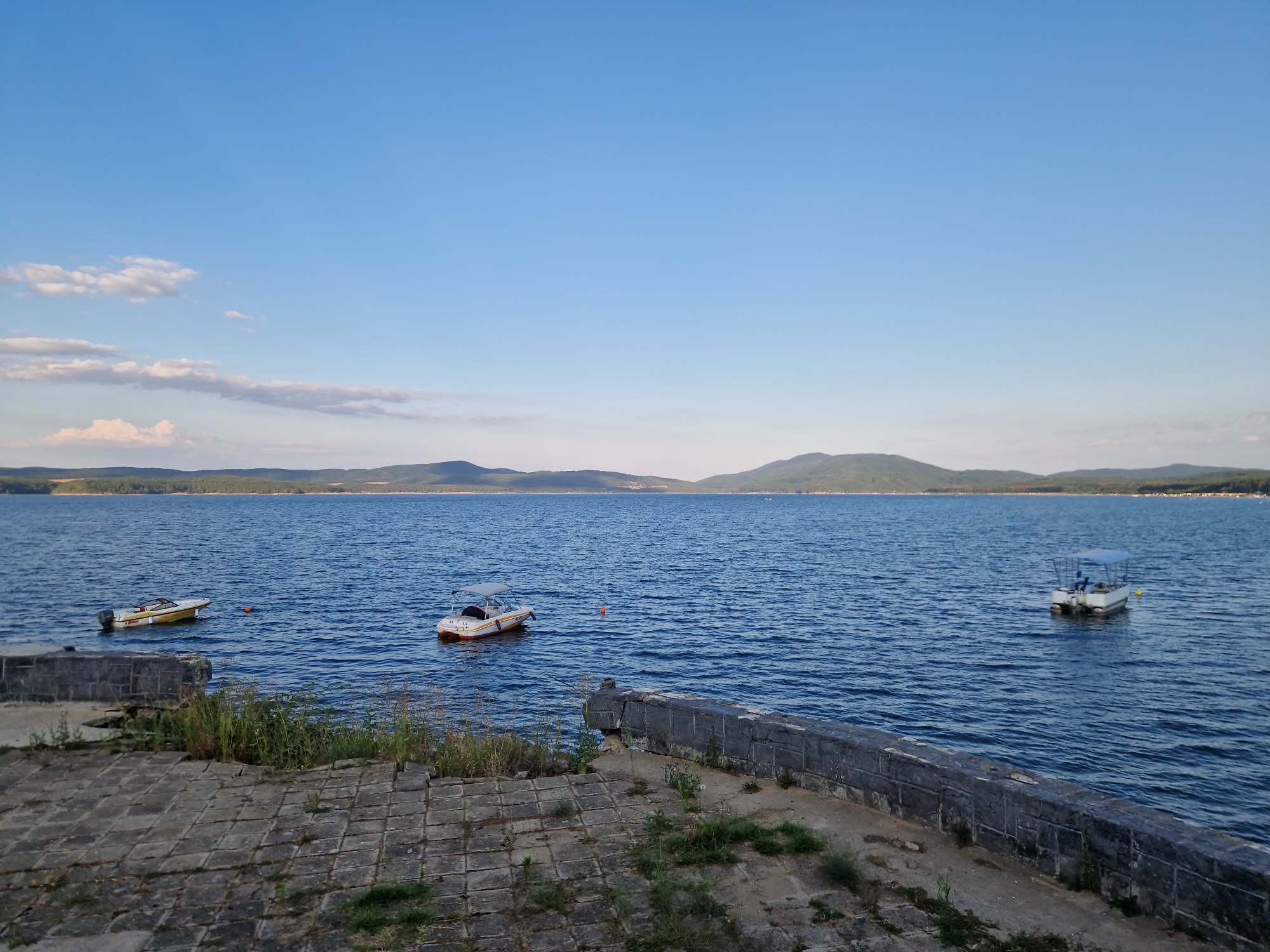 The image shows a wide, tranquil lake under a clear blue sky with a few scattered clouds. In the foreground, a paved area made of stone tiles slopes down towards the water. A rough, grey stone wall borders the lake on the right side of the image, with some sparse vegetation growing around its base.
The lake itself is calm, with gentle ripples on its surface. Three small boats are visible: two are closer to the foreground on the left, and one is further out on the right. The boat on the far left is a light-colored speed boat with a yellow stripe. Next to it, slightly closer to the viewer and further out, is a white and orange boat with a canopy. On the right side of the image, a white boat with a blue canopy is positioned further into the lake. An orange buoy is floating in the water between the two boats on the left.
In the distance, a hazy, tree-covered shoreline forms a gentle curve across the horizon, beyond which lie low, rolling hills or mountains. The overall impression is one of a peaceful, natural landscape. The scene is likely the Iskar Dam near the village of Dolni Pasarel, Bulgaria, a popular recreational area. There are no people visible in the image.