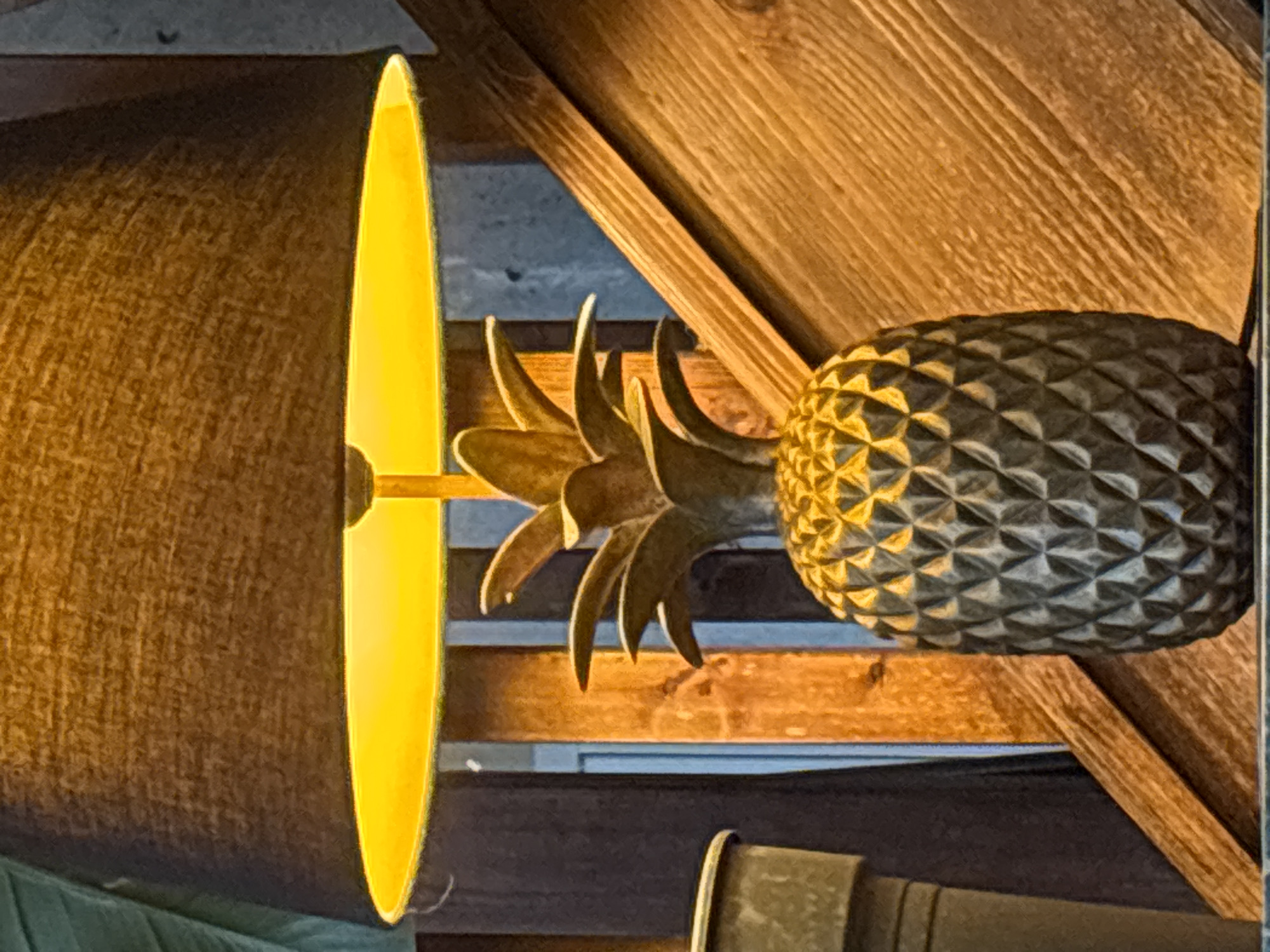 The image shows a pineapple-shaped lamp affixed to a wooden structure. The lamp features a textured, diamond-patterned pineapple body and stylized metal leaves around its base. A cylindrical lampshade is attached to the lamp, with a bright, narrow strip of light visible on its edge. The wooden structure behind the lamp appears to be part of an interior architectural element, possibly a beam or rafter. The background includes indistinct objects and surfaces. The image does not contain any information about London or the United Kingdom.