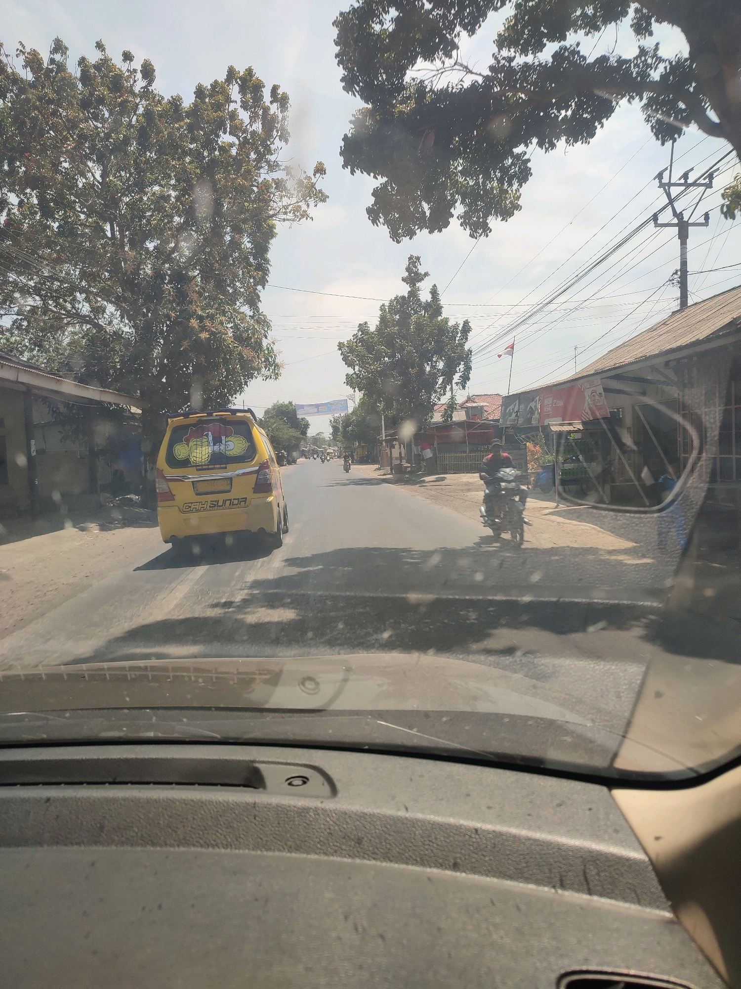 A low-angle, sunlit, outdoor, forward-facing view from inside a vehicle looking out through the windshield. The scene depicts a street in Desa Kedungwungu, Indonesia, with buildings and trees lining the asphalt road. A yellow minivan with a cartoon decal on the rear window is driving away from the camera in the middle of the road. To the right of the minivan, a person on a motorcycle is riding in the same direction. The sky is bright blue with some clouds, and electrical wires are visible overhead. The foreground shows the dusty interior of the vehicle, including part of the dashboard.