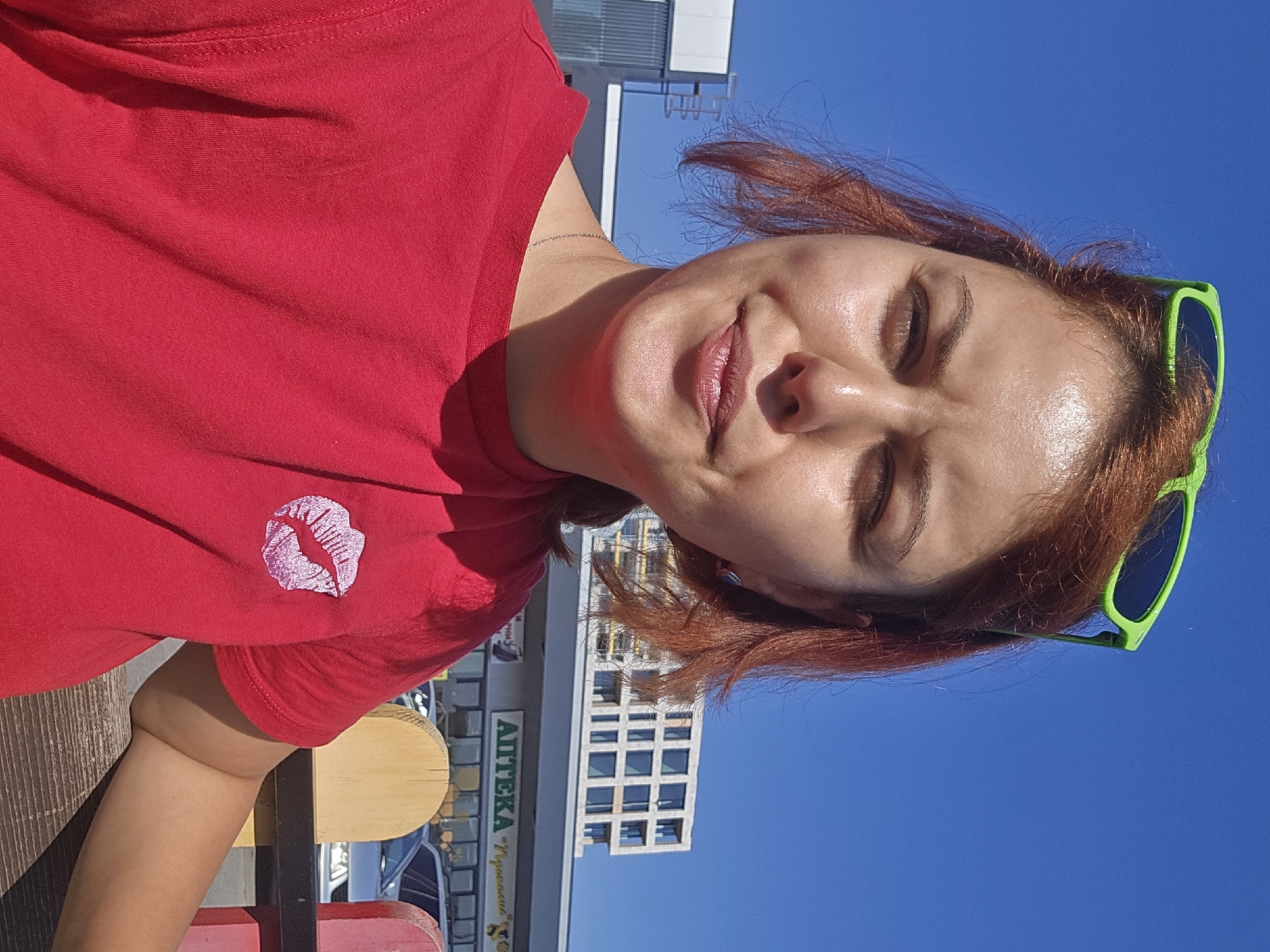 A woman with reddish-brown hair, wearing bright green sunglasses pushed up onto her head and a red t-shirt with a pink lipstick kiss graphic on the left chest, poses for a selfie outdoors under a clear blue sky. Her eyes are narrowed, and her lips are slightly pursed. In the background, a modern building with many windows and a sign reading "АПТЕКА" (Apteka, meaning Pharmacy) are visible, suggesting an urban setting. Part of a wooden fence or bench is in the foreground.
