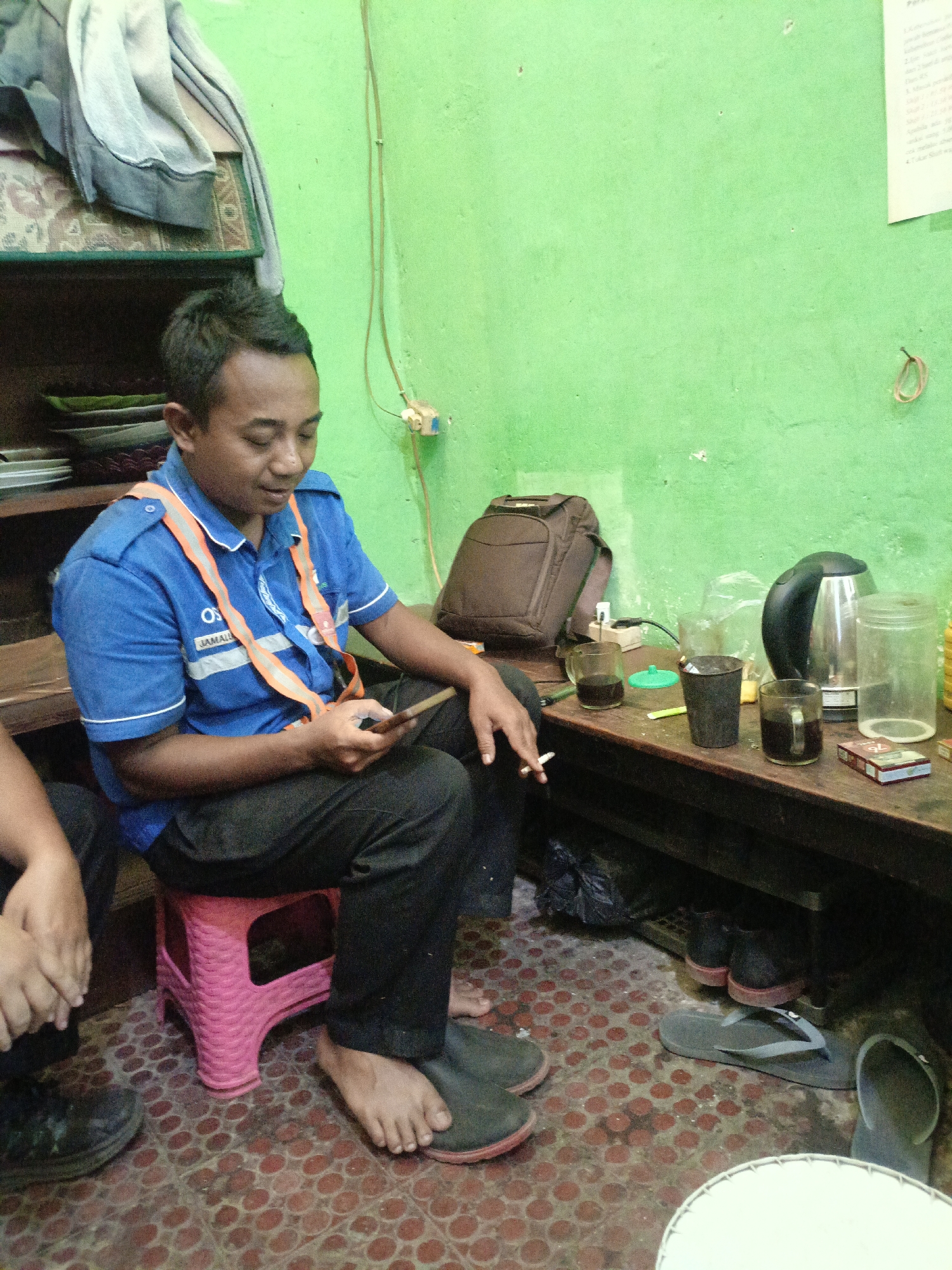 The image depicts an indoor scene in Kidul Dalem, Indonesia, likely a casual gathering place. A man wearing a blue uniform shirt with orange reflective stripes sits on a pink plastic stool, looking down at his mobile phone and holding a cigarette. He is barefoot and wearing dark pants. Beside him, another person's legs and hands are visible, suggesting they are also present. A wooden table in the background holds several items, including glasses of dark liquid, a black electric kettle, a clear plastic jar, and a pack of cigarettes. A brown backpack rests on the table, and a shelf above holds various objects, including what appears to be clothing. The walls are painted a vibrant green. The floor is tiled with a red and brown pattern.