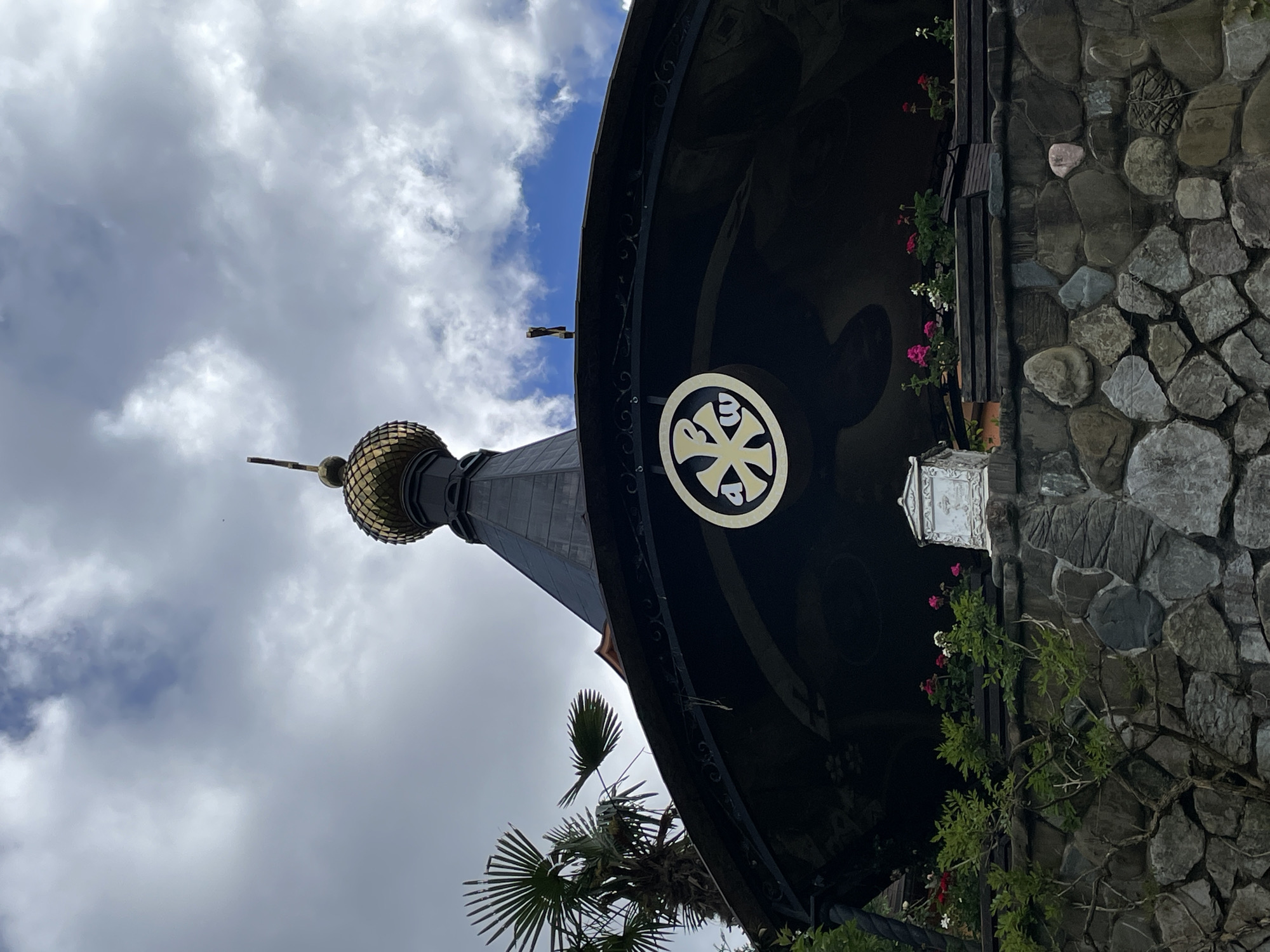 Captured from a low angle, the image focuses on architectural details against a backdrop of sky with scattered clouds. A building, characterized by a rounded, dark-colored awning adorned with a circular logo, features a tower with a golden, patterned dome. A stone wall stands adjacent to the awning. Vegetation, including flowers and vines, is visible. The image is taken in Solokh-Aul, Russia.