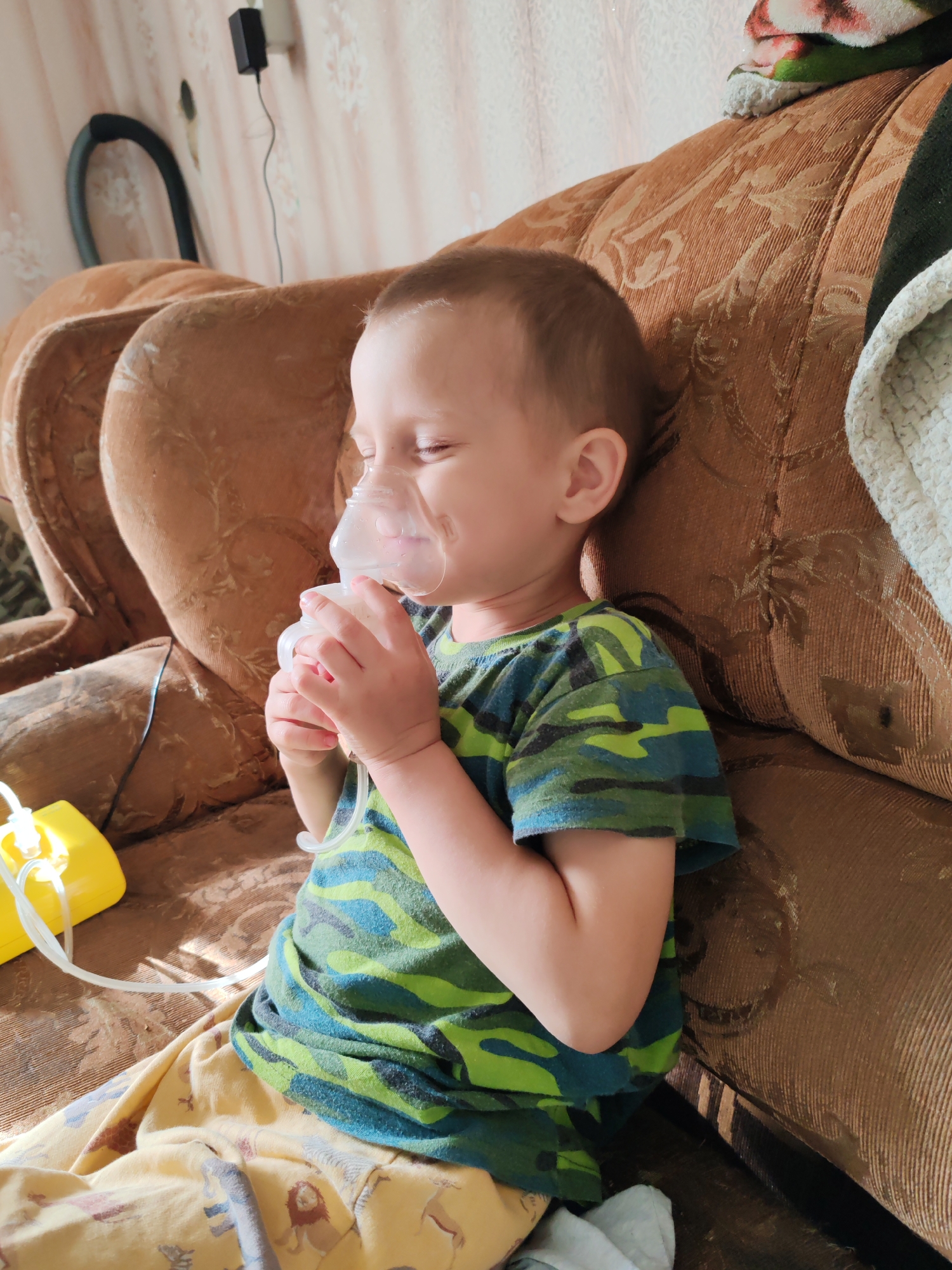 A young child is seated on a brown upholstered couch with a nebulizer mask covering their mouth and nose. The child is wearing a green and blue camo-patterned shirt and yellow pants with animal prints. Their eyes are closed. The nebulizer machine, which is yellow, is visible on the couch next to the child. In the background, parts of the room are visible, including the wall and a section of what appears to be a vacuum cleaner hose. The location is Чухлэм, Russia.