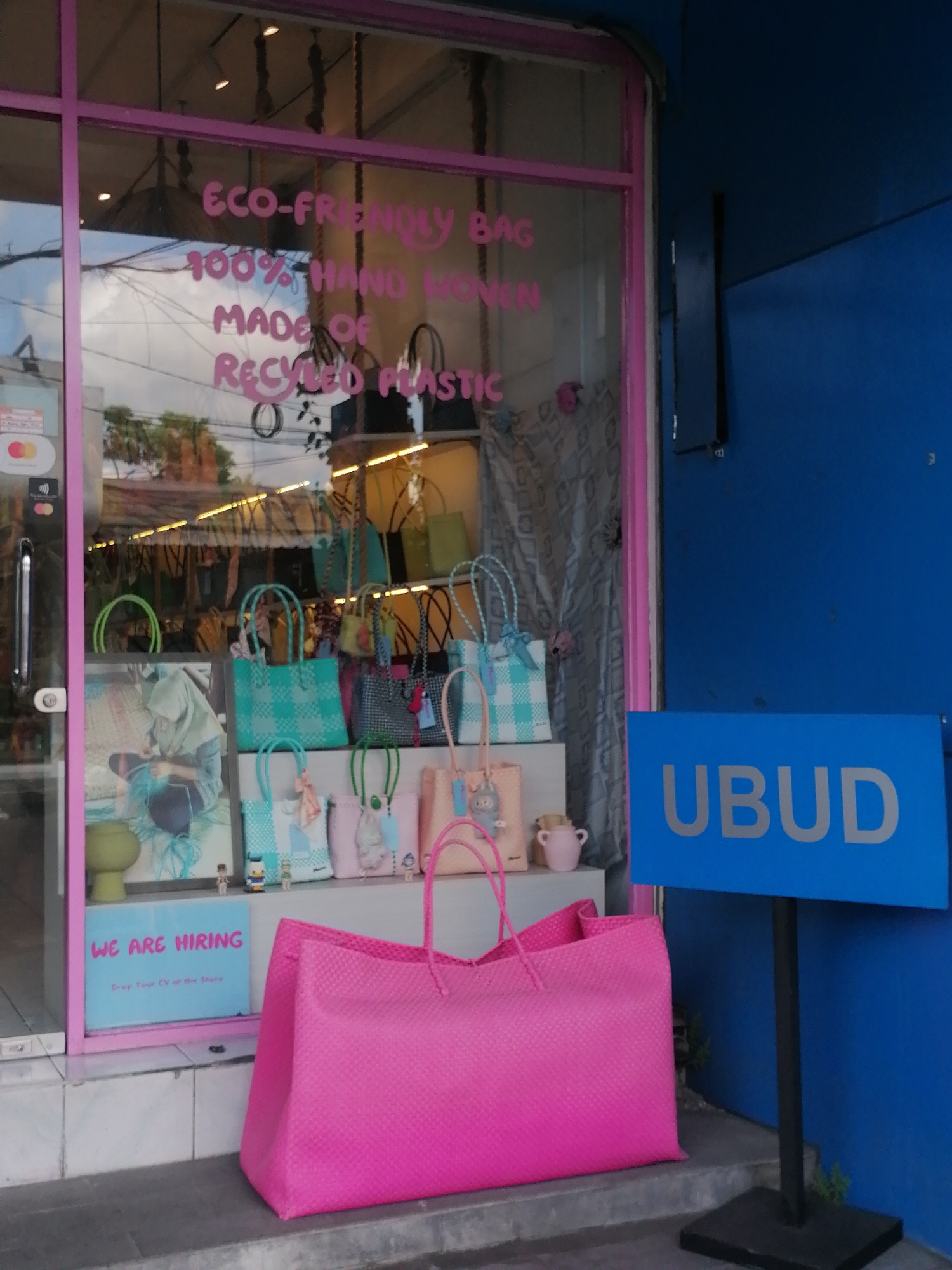 The image shows the exterior of a store in Ubud District, Indonesia. The store has a large glass window with pink trim. Inside the window, various woven bags are displayed on shelves. Text on the window reads "ECO-FRIENDLY BAG 100% HAND WOVEN MADE OF RECYCLED PLASTIC". Below this text, there is a blue sign with white text that says "UBUD". To the left of the sign, another blue sign reads "WE ARE HIRING Drop Your CV at the Store". In front of the store, a very large bright pink woven bag sits on the ground. The store appears to be on a street with a blue wall to the right of the entrance.