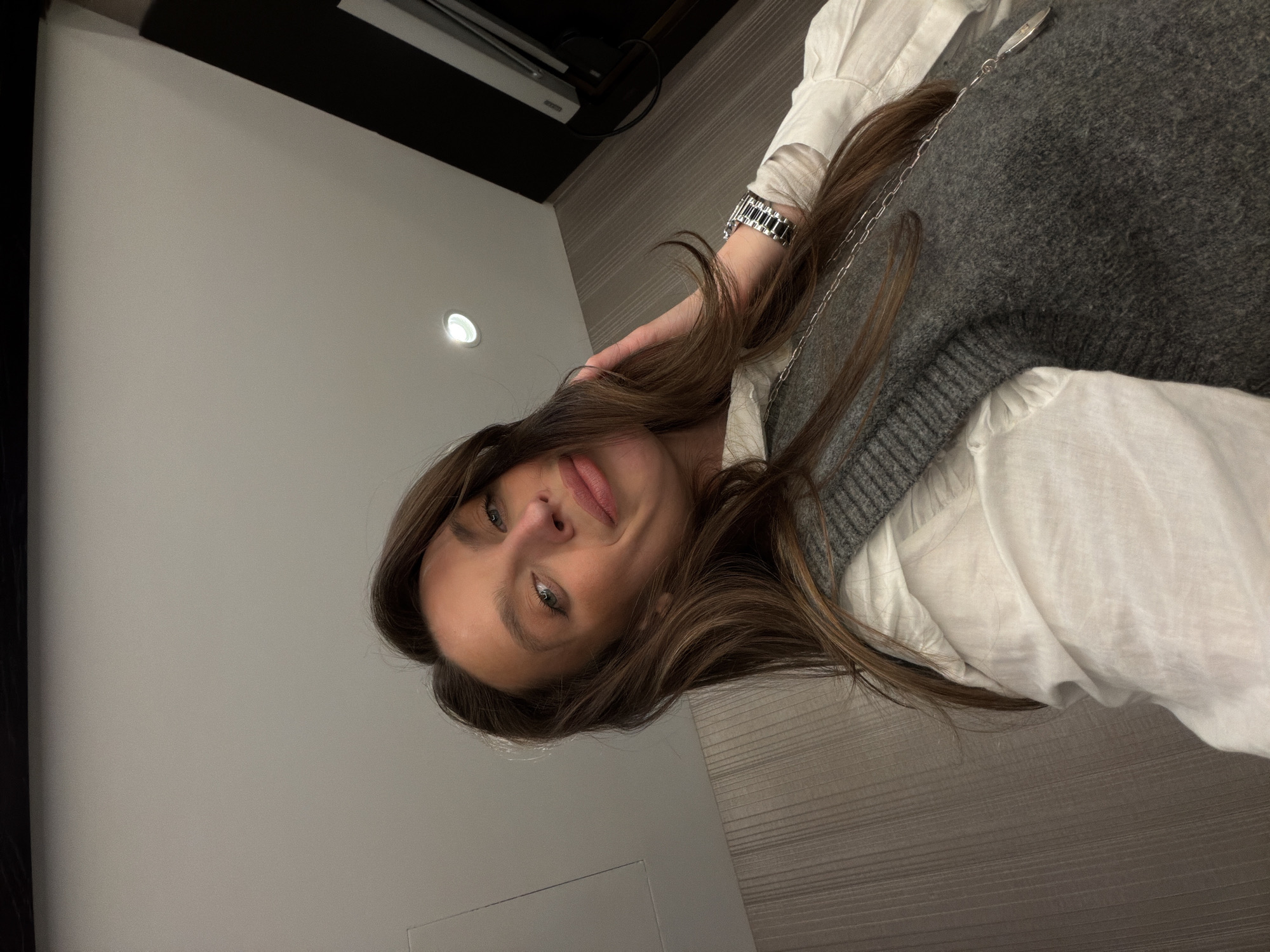 A selfie shows a woman with long brown hair, fair skin, and green eyes. She is wearing a white shirt with a gray knit vest over it. On her left wrist is a silver watch. The background consists of a white wall with a recessed light, a black panel above it, and a light-colored wooden surface. The location, Budva, Montenegro, cannot be confirmed or refuted with the visual evidence.