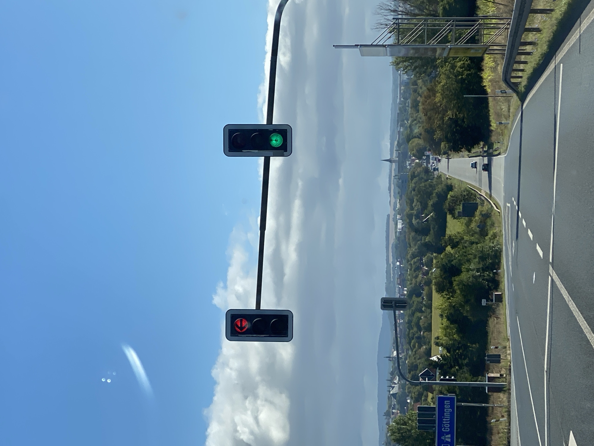A long-range outdoor shot captures a highway leading towards a town with a church spire, identified as likely being in the vicinity of Werther, Germany, given the "Göttingen" sign visible. The sky is bright blue with scattered white clouds, and a faint white contrail or atmospheric anomaly is visible in the upper left. Two traffic lights are prominent in the foreground, one showing a red left arrow and the other displaying a green light. The road is paved and marked with white lines. Trees and fields flank the road, and several cars are visible on the highway in the distance. A metal guardrail runs along the right side of the road, and a metal pole with signage is present on the right.