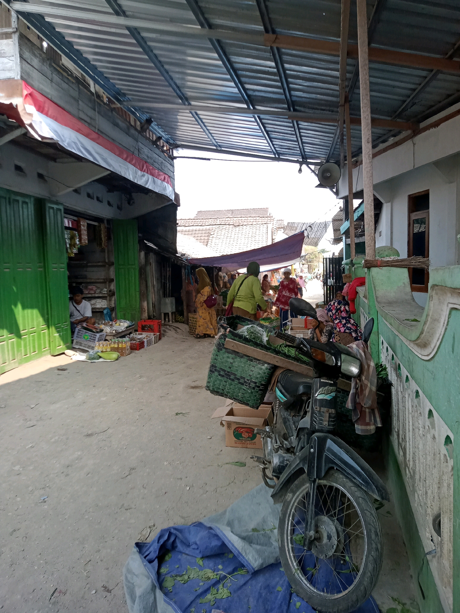 The image depicts a street scene in Tanggungharjo, Indonesia, likely a marketplace. A corrugated metal roof supported by metal poles covers a portion of the street. In the foreground, a black motorcycle is parked, with green baskets containing produce on its rear. A blue tarp with green leaves is spread on the ground in front of the motorcycle.
To the left, a storefront with green folding doors is visible. A young man sits inside the store, surrounded by crates of goods. Produce, including gourds and bottles of liquid, is displayed on the ground in front of the store. Further down the street, several women in traditional clothing are seen browsing or selling goods under a purple canopy.
The ground is dirt and gravel. In the background, buildings with tiled roofs are visible. A large Indonesian flag, red and white, is draped along the top of one of the structures on the left. The overall impression is of a bustling local market on a sunny day.