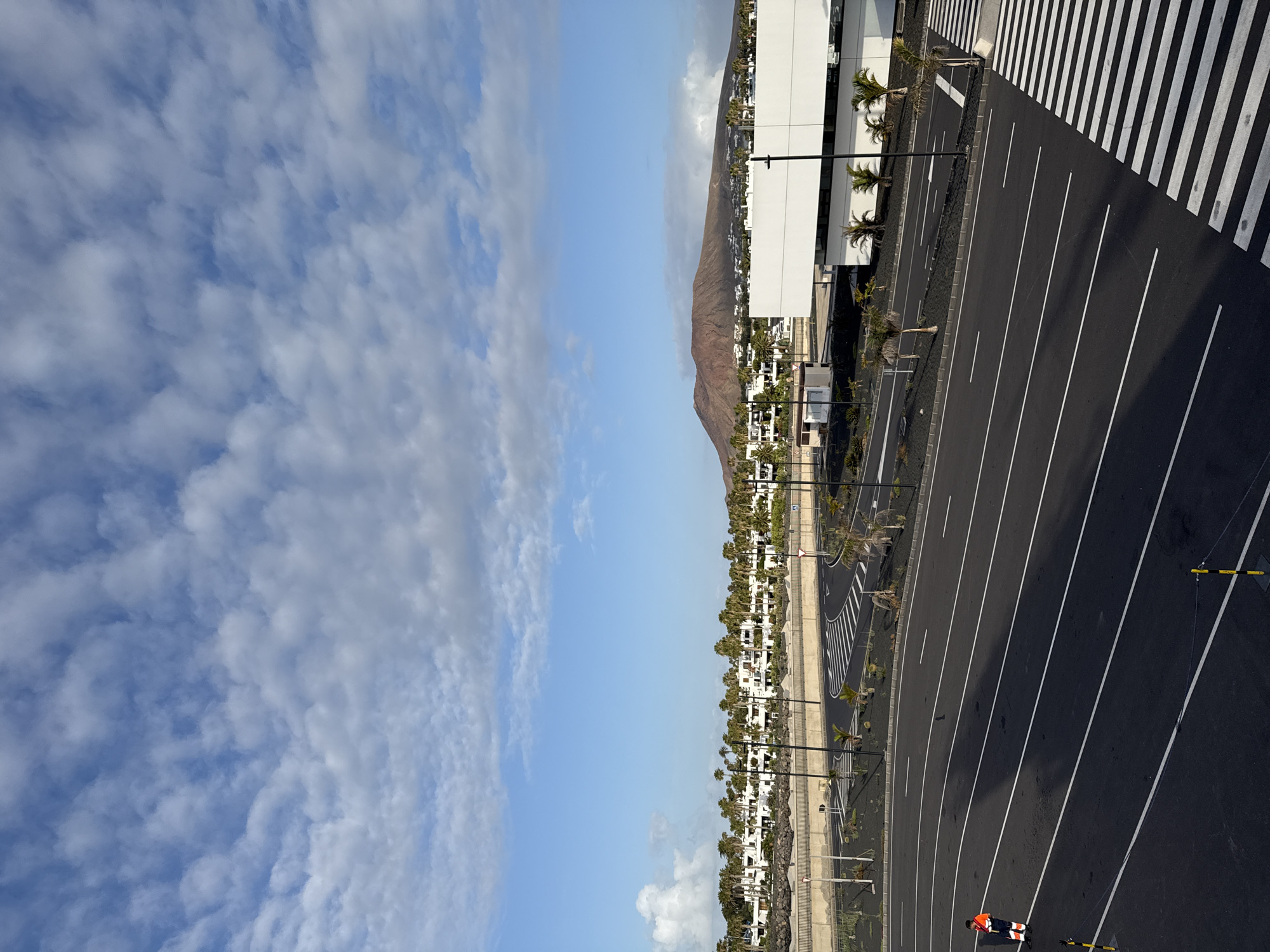 The image shows a view over Yaiza, Spain, a municipality in the Canary Islands. A brown-colored mountain rises in the background. The sky is blue with some scattered clouds. A dense row of white buildings stretches across the midground, interspersed with trees. The foreground consists of an asphalt parking lot with white painted lines, a crosswalk, and utility poles. An individual wearing a safety jacket can be seen in the lower-right corner of the image.