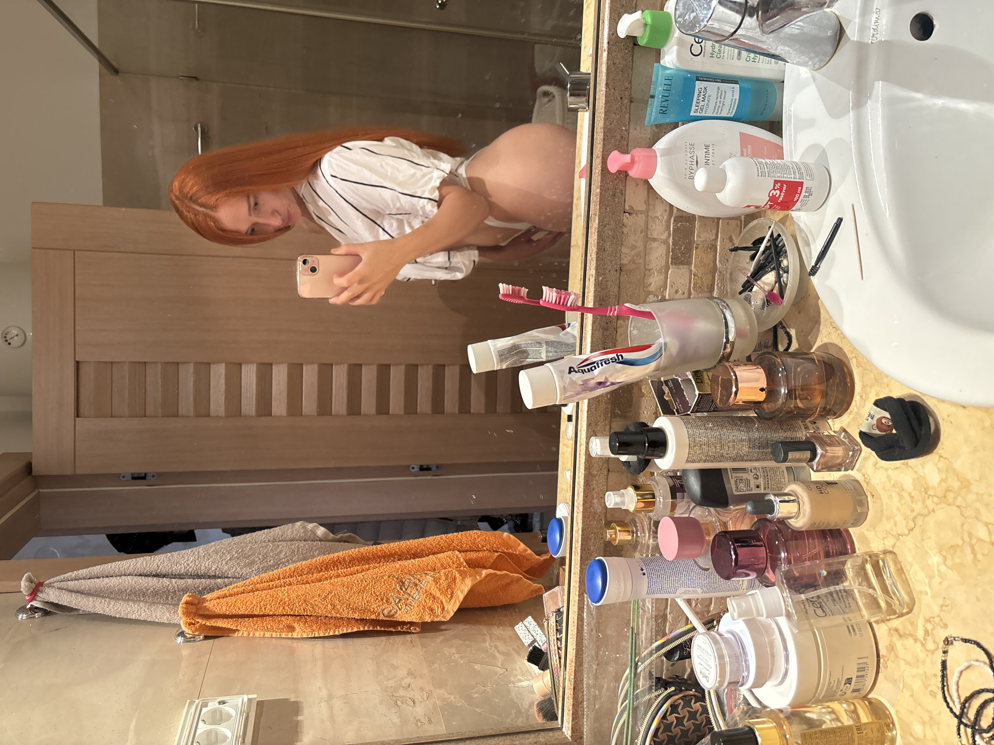 A young woman with long, bright red hair stands in a bathroom, taking a selfie in a mirror. She is wearing a white shirt with black pinstripes and white underwear. Her body is partially visible in the reflection, showing her profile from the side. The bathroom is well-lit and features a marble countertop with an assortment of toiletries, including bottles of skincare products, perfumes, a tube of toothpaste, and toothbrushes. Two towels, one orange and one beige, hang on hooks on the wall. The overall impression is of a personal, private space. The scene is consistent with a bathroom located in Budva, Montenegro, known for its coastal beauty and resort-style accommodations.