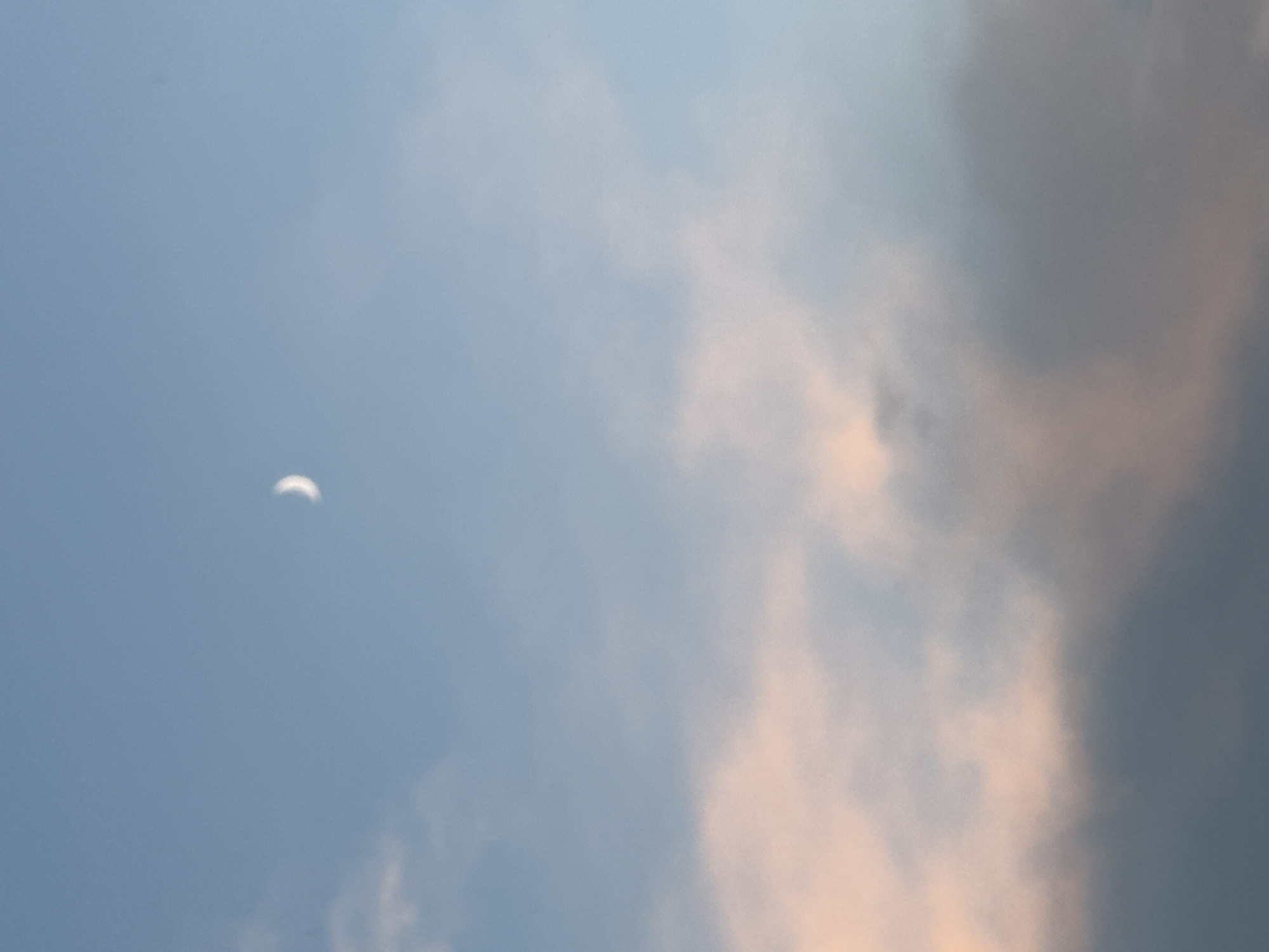 The image depicts a pale crescent moon visible in a pale blue sky. Below the moon are soft, diffused clouds, some tinged with a peachy or pink hue, indicating either sunset or sunrise. The overall scene is tranquil and atmospheric, suggesting an open sky view without any urban elements or identifiable landmarks. It is impossible to determine the specific location within Moscow, Russia, from the provided image.