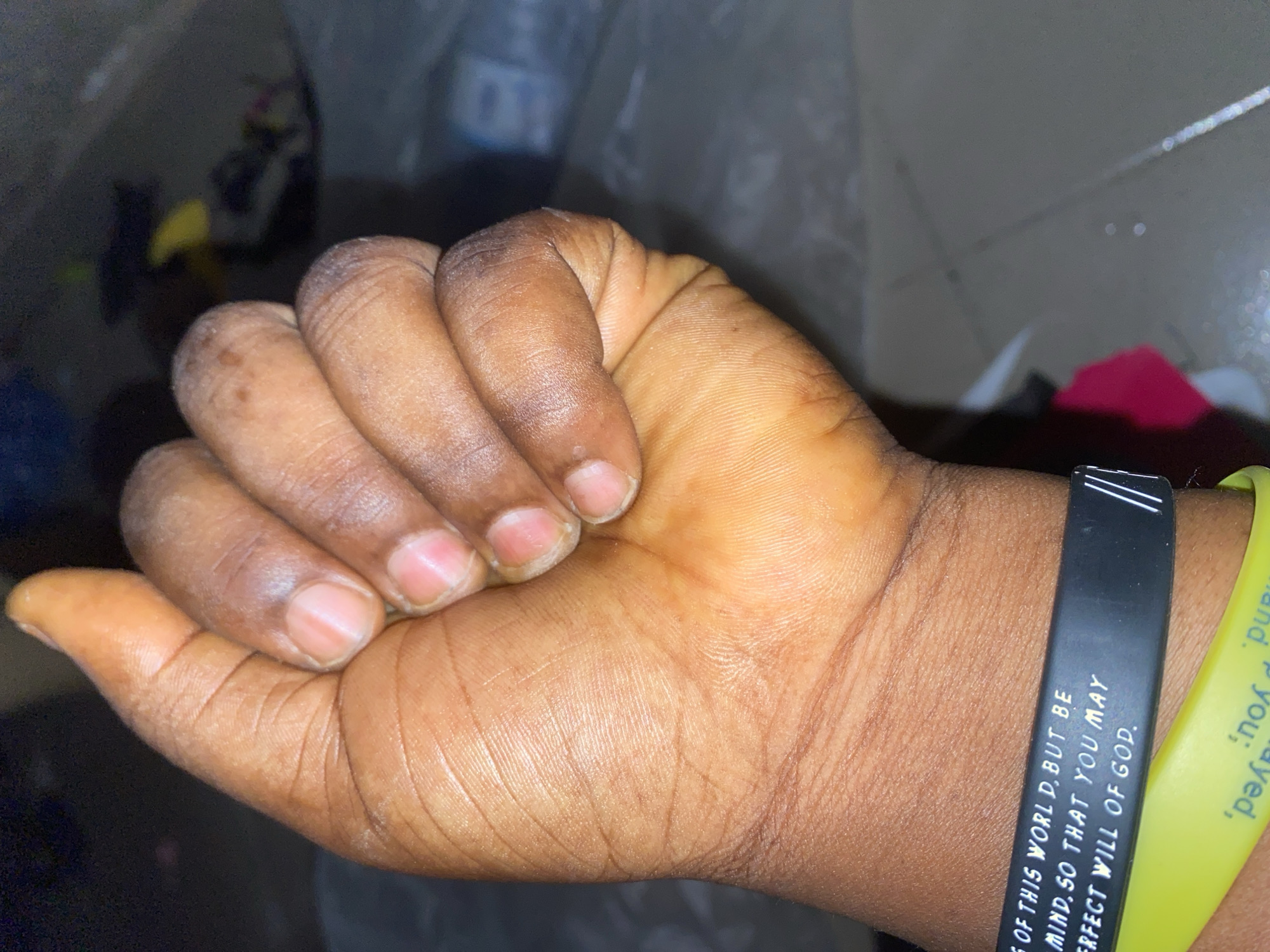 The image shows a close-up of a closed fist with fingernails visible. The skin is dark brown and appears to be well-moisturized. There are several wrinkles and lines on the palm and fingers, indicating age or frequent use. Two wristbands are visible on the wrist. The upper wristband is black and has white text that reads "OF THIS WORLD, BUT BE MIND, SO THAT YOU MAY PERFECT WILL OF GOD." The lower wristband is yellow and has white text that is partially visible, appearing to say "'Hand,' you prayed". The background is dark and out of focus, with hints of other objects and possibly clothing. The scene appears to be indoors, with a tiled floor visible in the background. The lighting is dim, with some light reflecting off the surfaces.