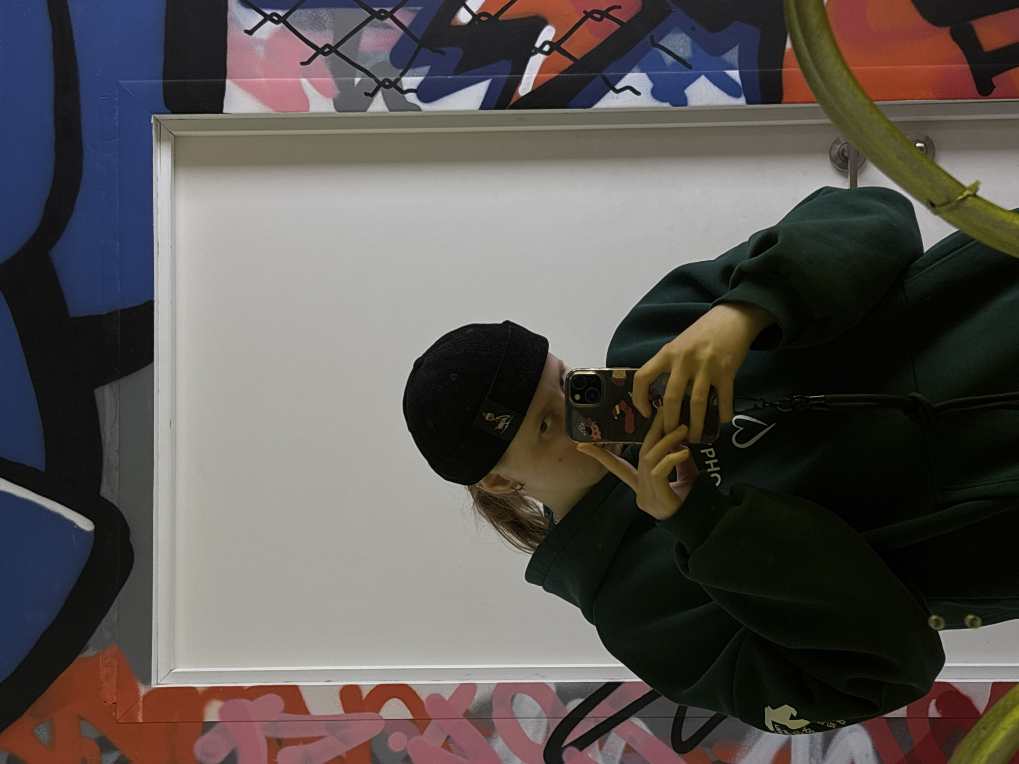 The image depicts a young person taking a mirror selfie in an urban art environment. The individual, wearing a dark green hoodie and a black beanie, holds a smartphone with a decorated case, capturing their reflection in the mirror. Their face is partially obscured by the phone.
The mirror itself frames a plain white wall, but its border is surrounded by a wall extensively covered in vibrant, abstract graffiti art. The graffiti features a dynamic mix of blue, black, red, orange, pink, grey, and white spray paint, with a black chain-link fence motif drawn over some of the patterns at the top edge. On the right side of the image, a curved, yellow-green object, possibly a pipe or a structural element, is visible.
Details on the person's attire include a white heart graphic and partial text reading "PHO" on the chest of their hoodie. A small logo is also present on the black beanie. The setting suggests an indoor creative space or gallery, possibly in Perm, Russia, given the art style and prominent graffiti. The lighting appears to be artificial, typical of an indoor environment. No specific time of day or emotional state is discernible beyond the casual act of taking a selfie.